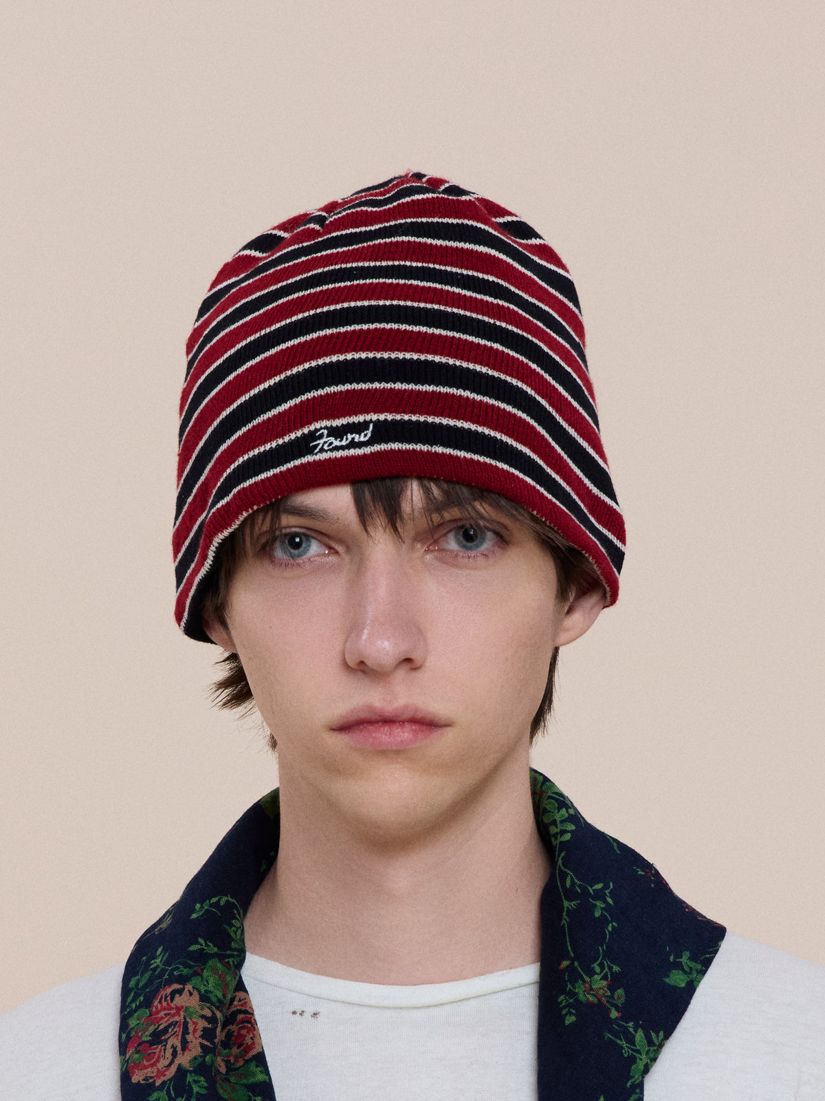 A young person with straight brown hair wears the Stripe Logo Beanie by FOUND and a patterned shirt, facing forward with a neutral expression against a plain background.