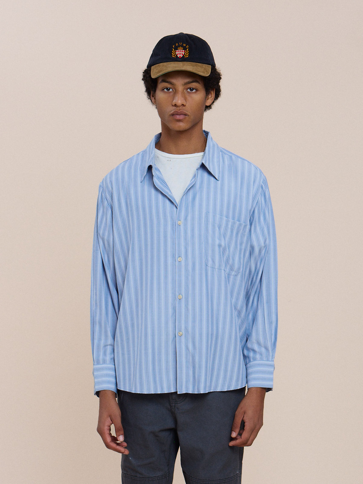 Model wears the FOUND Stripe Pocket Oxford Shirt, unisex relaxed fit, layered over a white tee and dark pants against a light background.