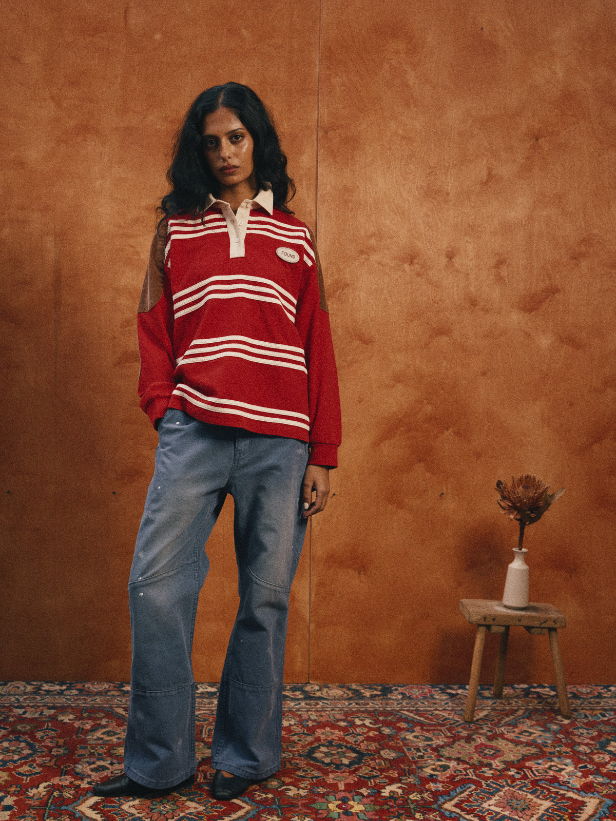 Woman stands indoors on a patterned rug, wearing the FOUND Striped Corduroy Rugby Polo and blue jeans, beside a wooden stool and dried flowers against a brown wall.