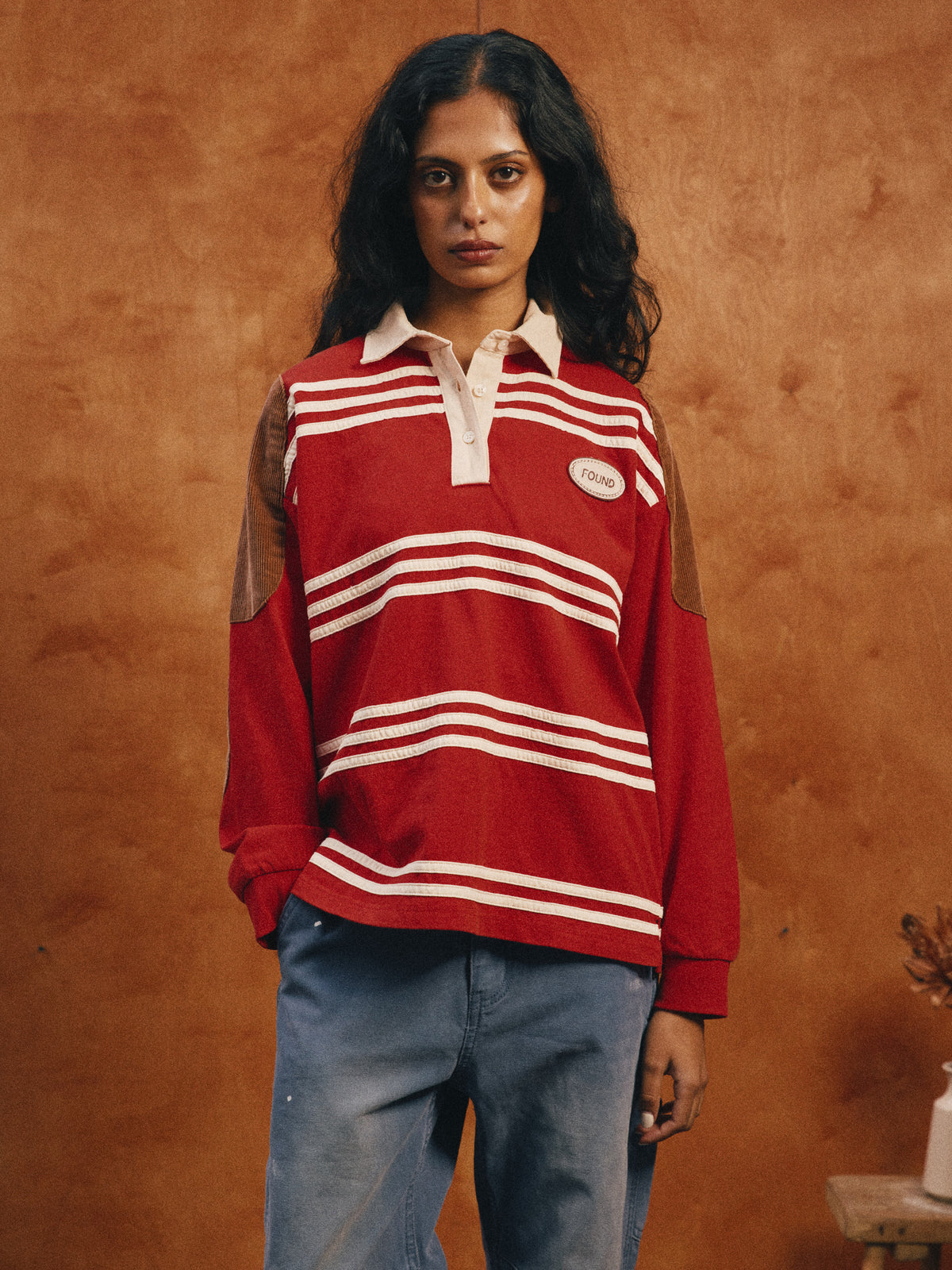 A woman with long dark hair wears the FOUND Striped Corduroy Rugby Polo and blue jeans, standing against a brown textured background.