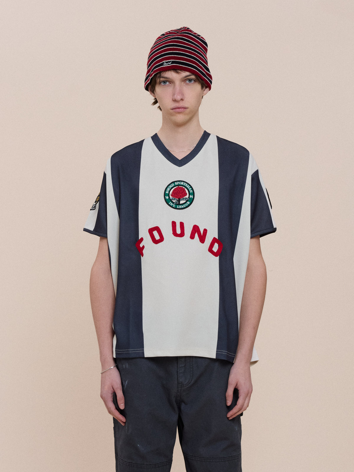 Person wearing a red-striped beanie, FOUND Striped Stateman Jersey with badge, and dark shorts, standing against a plain beige background.