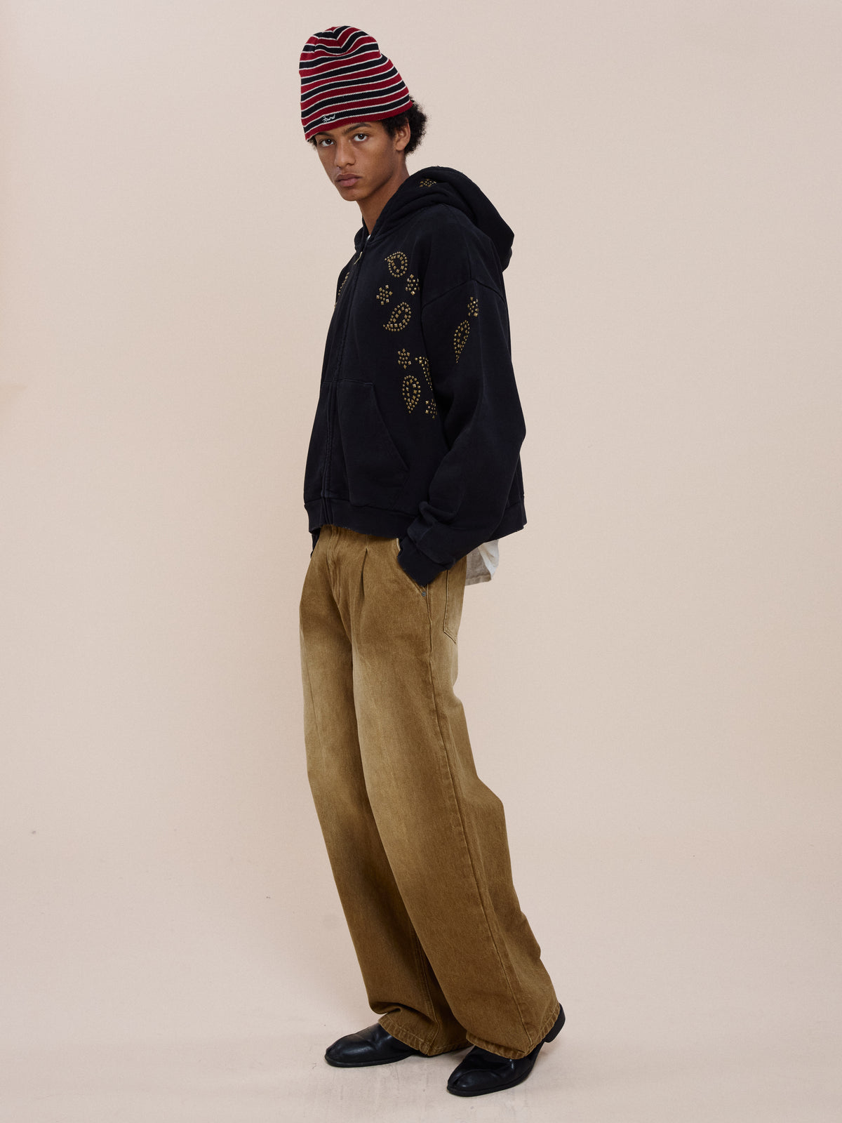 A person in the FOUND Studded Zip Up Hoodie, wide brown pants, black shoes, and a red-striped beanie stands against a plain light background.