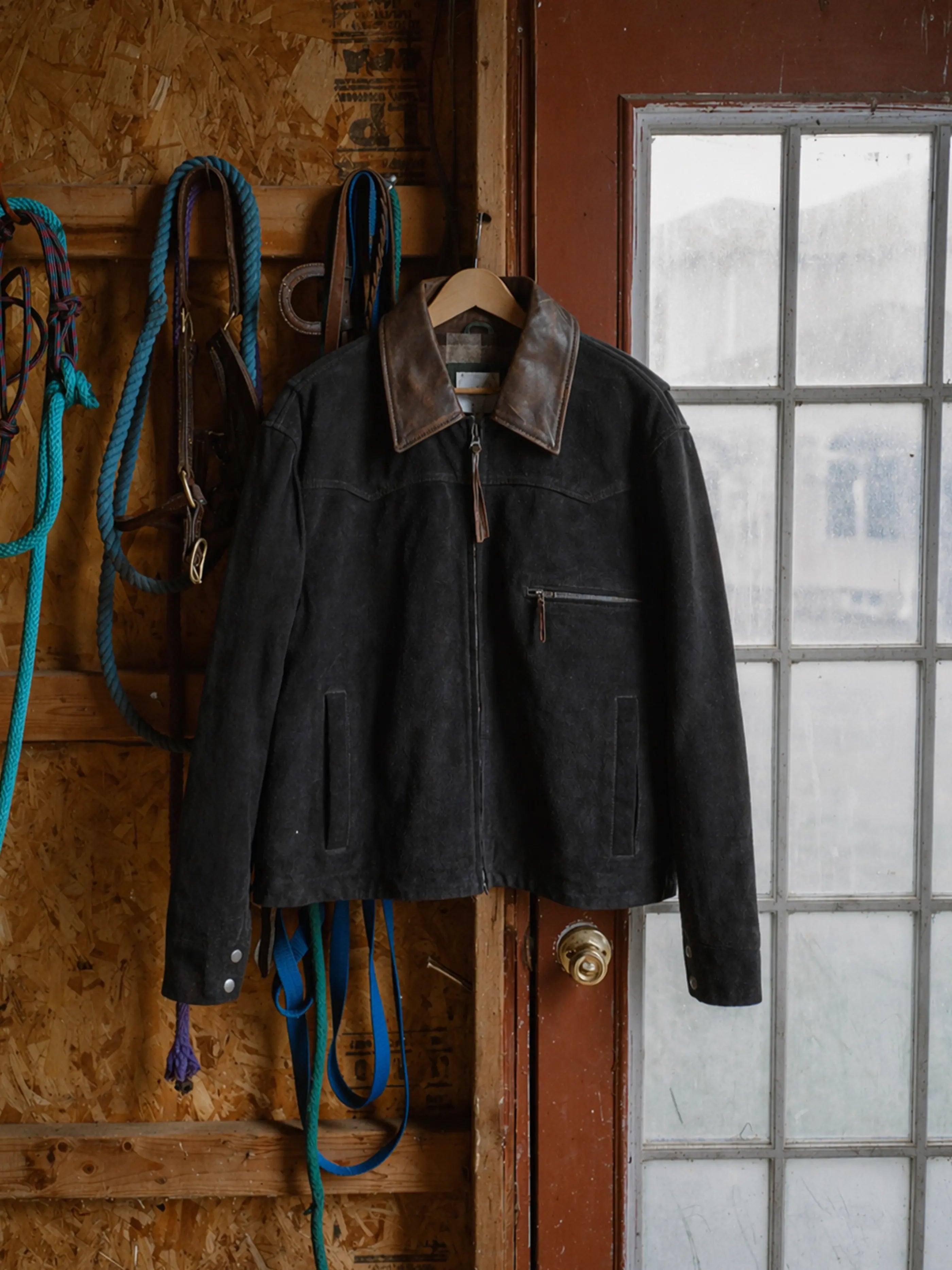 The FOUND Suede Western Work Jacket features black suede and a brown leather collar, hanging on a wooden hanger beside blue and green ropes in a rustic room with a glass door.