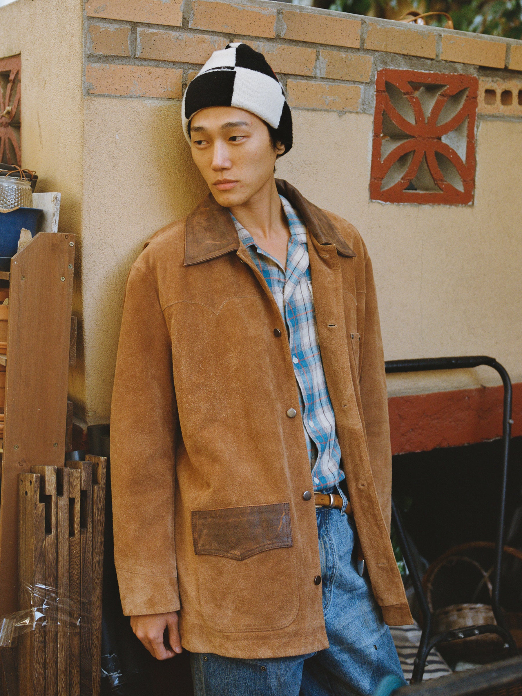 Wearing the FOUND Suede Contrast Chore Jacket, a plaid shirt, jeans, and a black-and-white beanie, a person stands against an outdoor stucco wall with decorative brickwork.