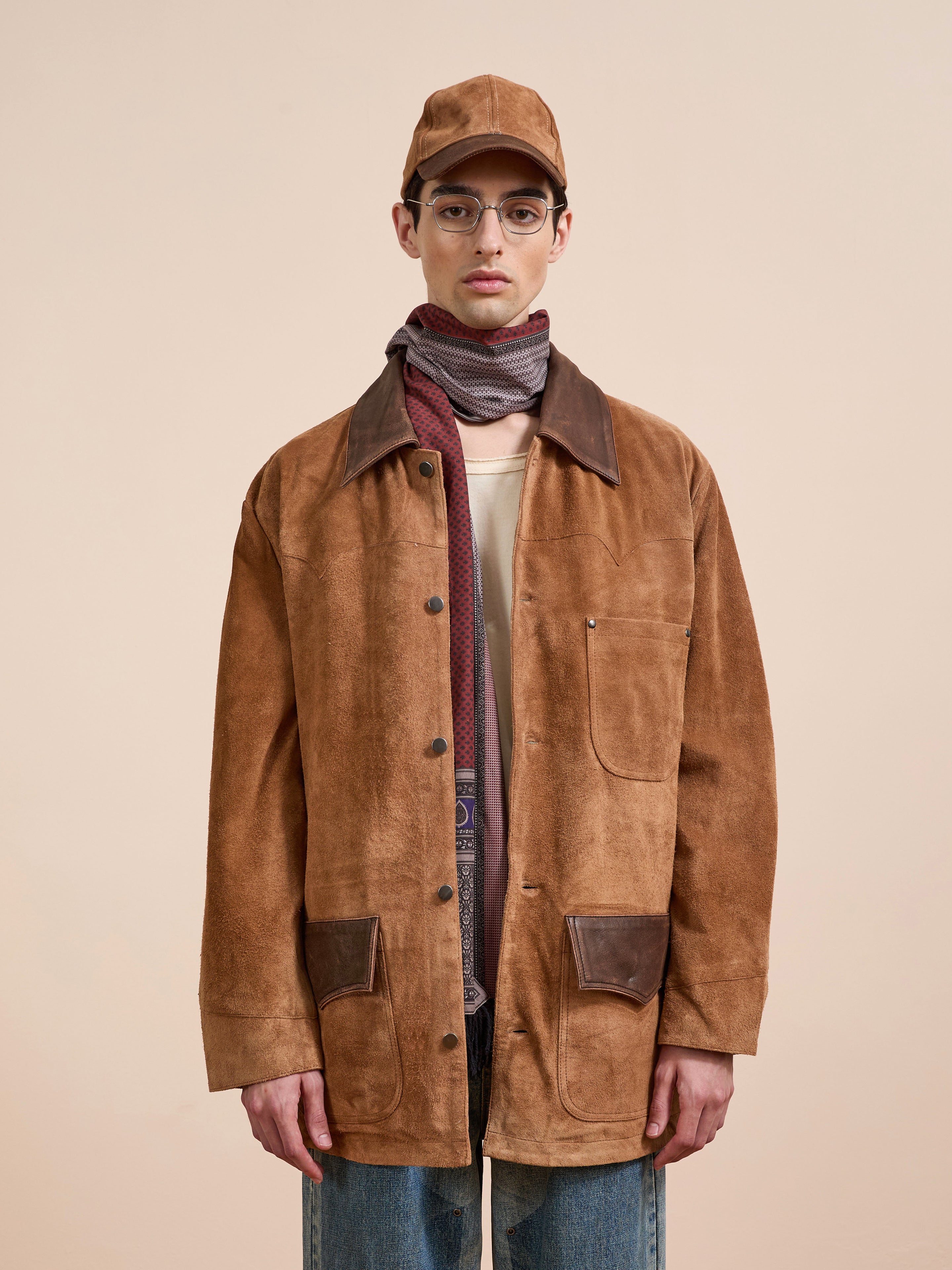 A man wearing the FOUND Suede Contrast Chore Jacket, a matching brown cap, glasses, striped scarf, and blue jeans stands against a neutral background.