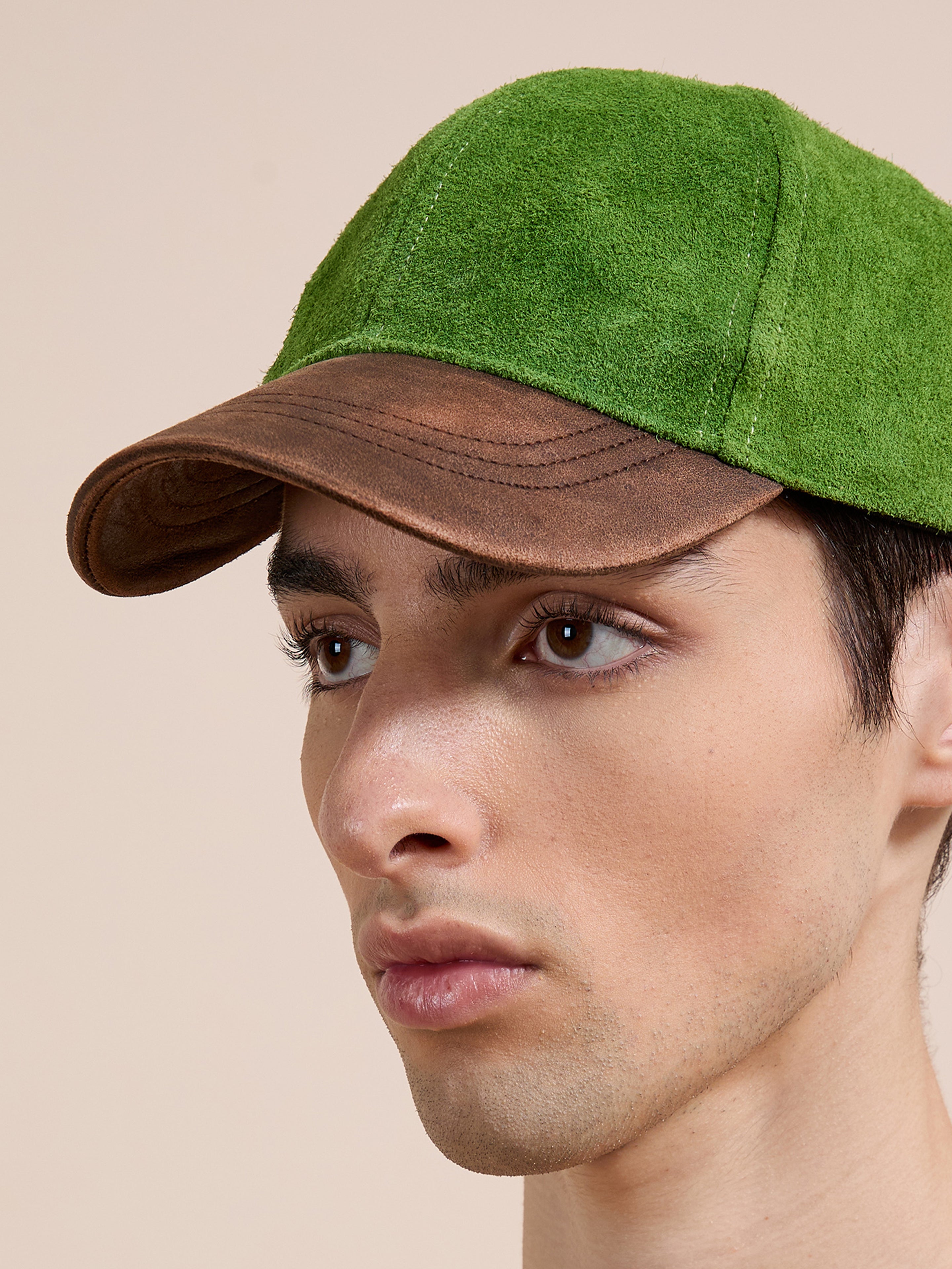 A person wears the Found Suede Leather Contrast Distressed Cap, featuring green suede and a brown distressed leather brim, while looking to the side against a neutral background.