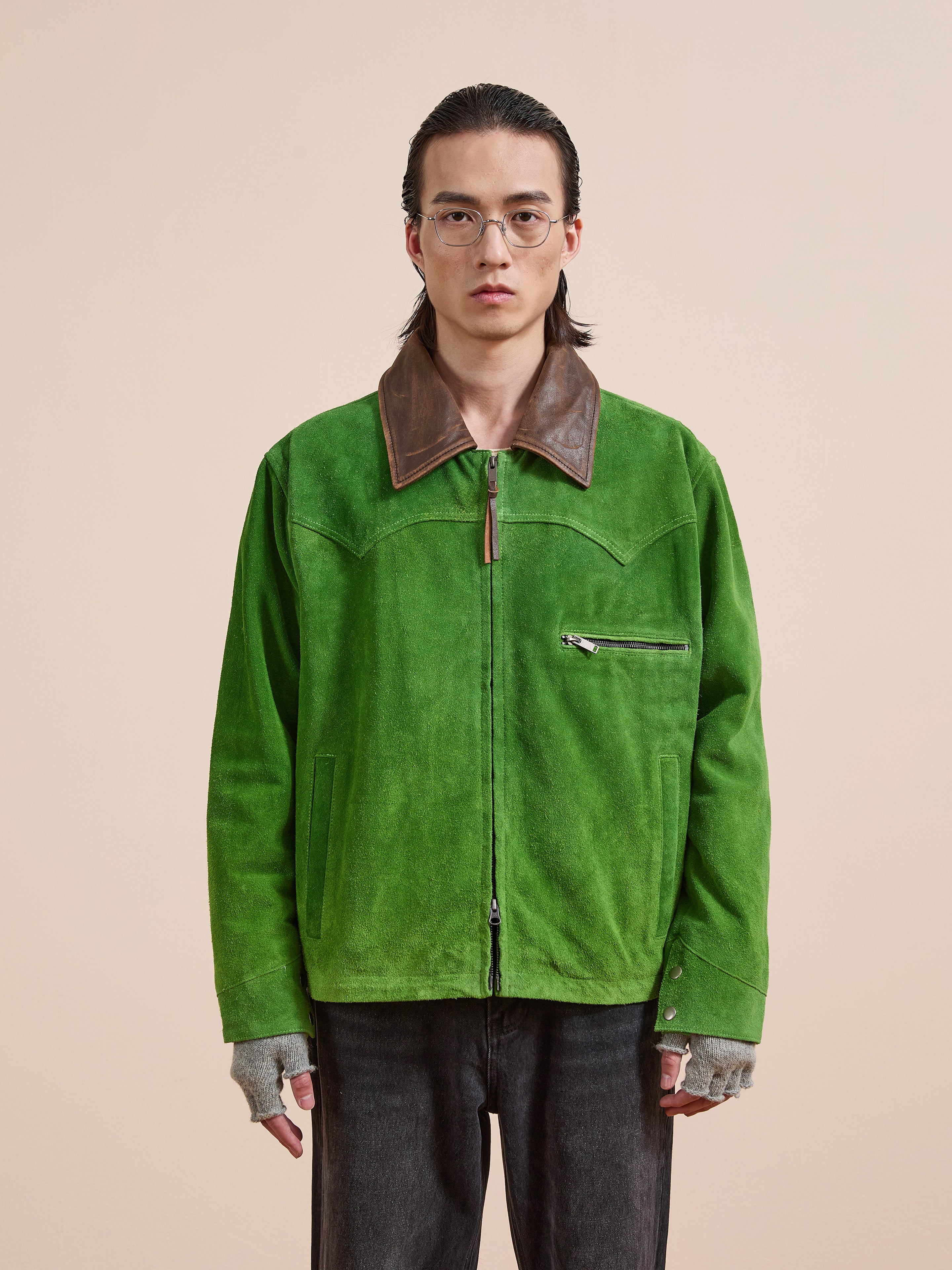 A person wearing glasses, the FOUND Suede Western Barn Jacket with a brown leather collar, fingerless gray gloves, and black jeans stands against a plain beige background.