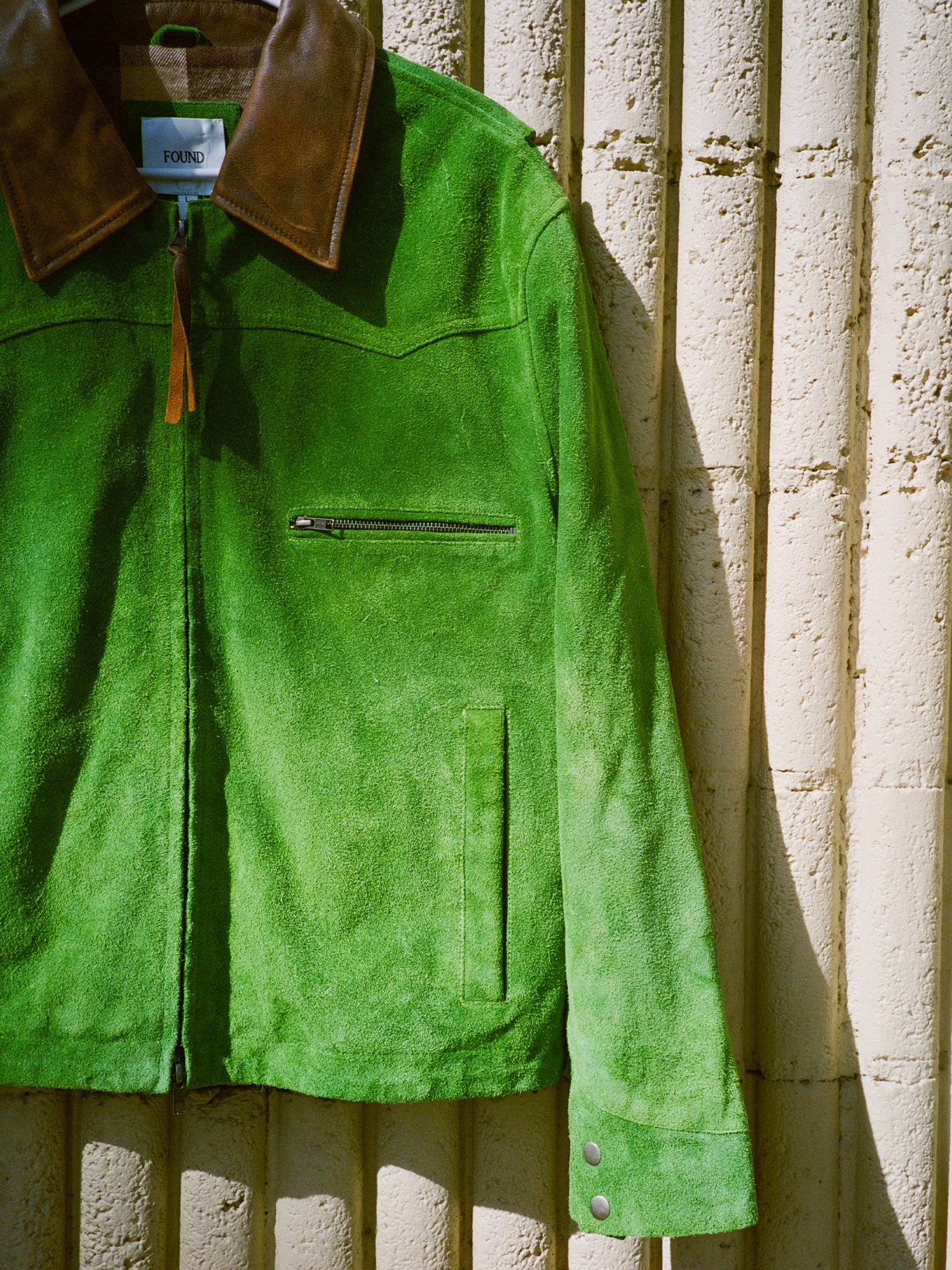 The FOUND Suede Western Barn Jacket in bright green suede, featuring a brown leather collar and zippered chest pocket, hangs against a textured beige wall.