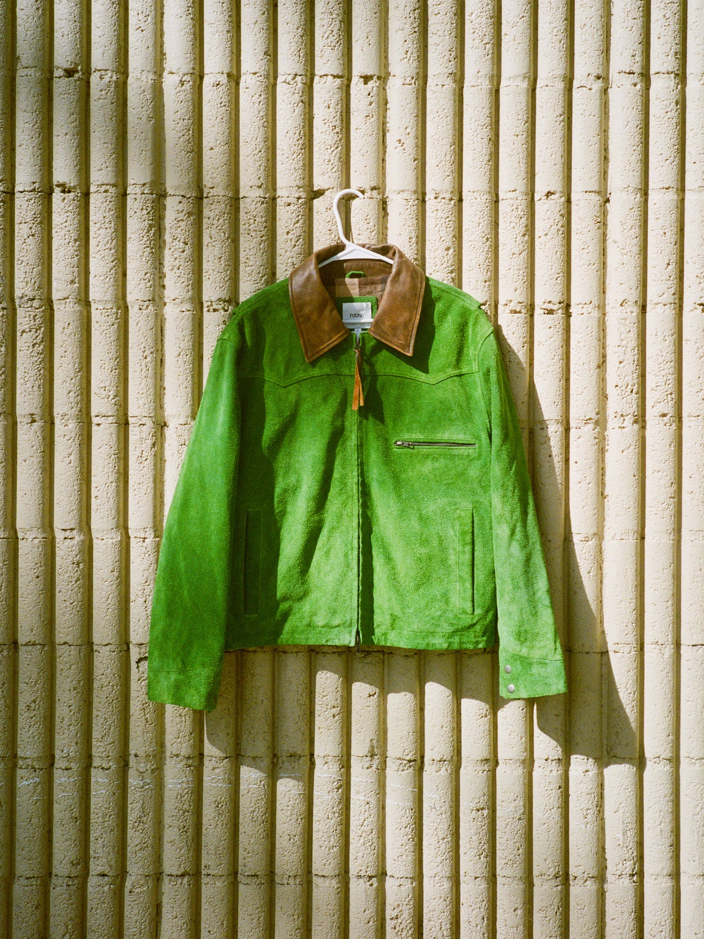 The FOUND Suede Western Barn Jacket, featuring a green suede body and brown collar, hangs on a white hanger against a textured beige concrete wall.