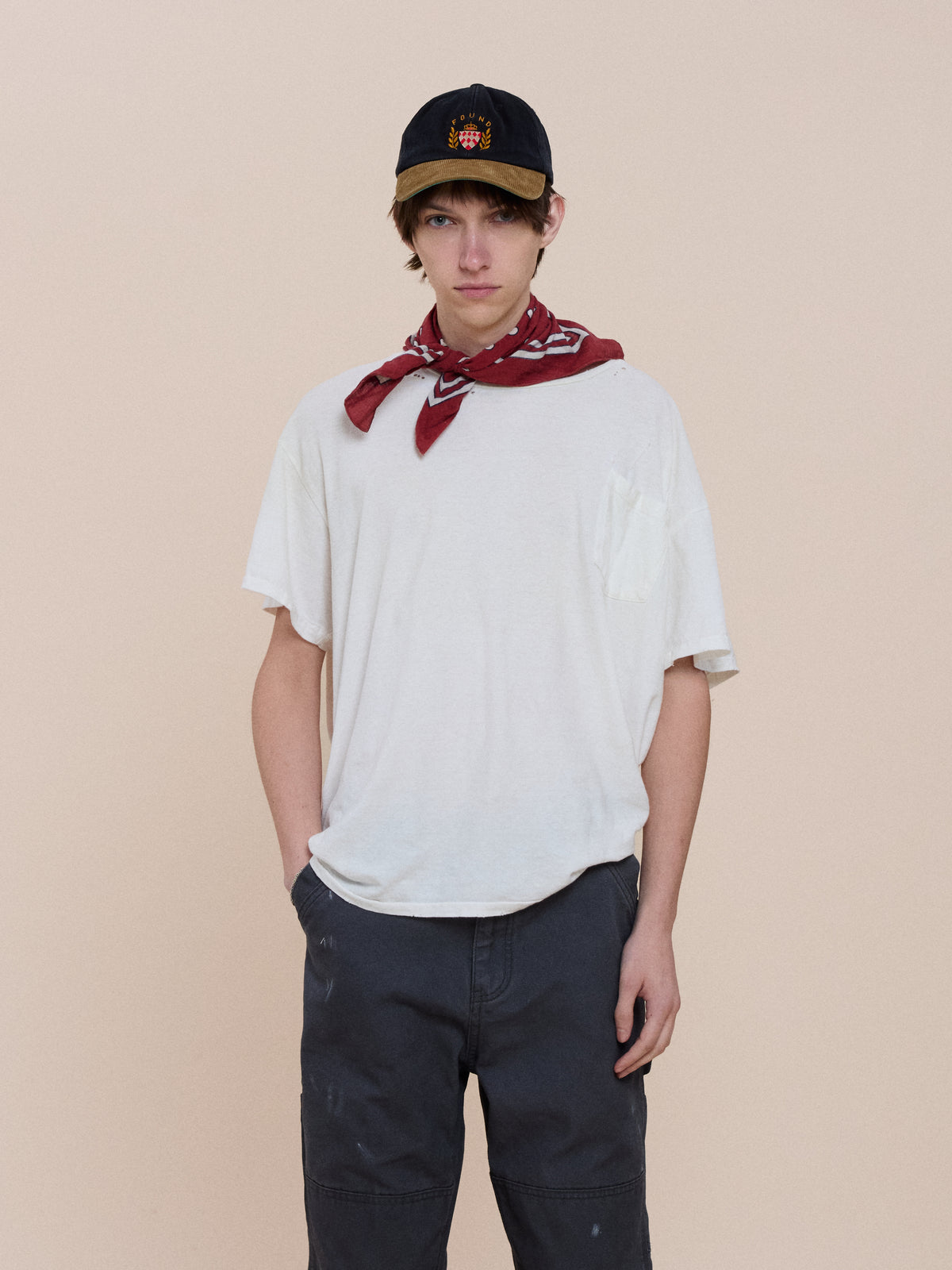 A young person in a black cap, red bandana, white tee, and FOUND Sunbleached Utility Pants stands against a plain light beige background with one hand in pocket.