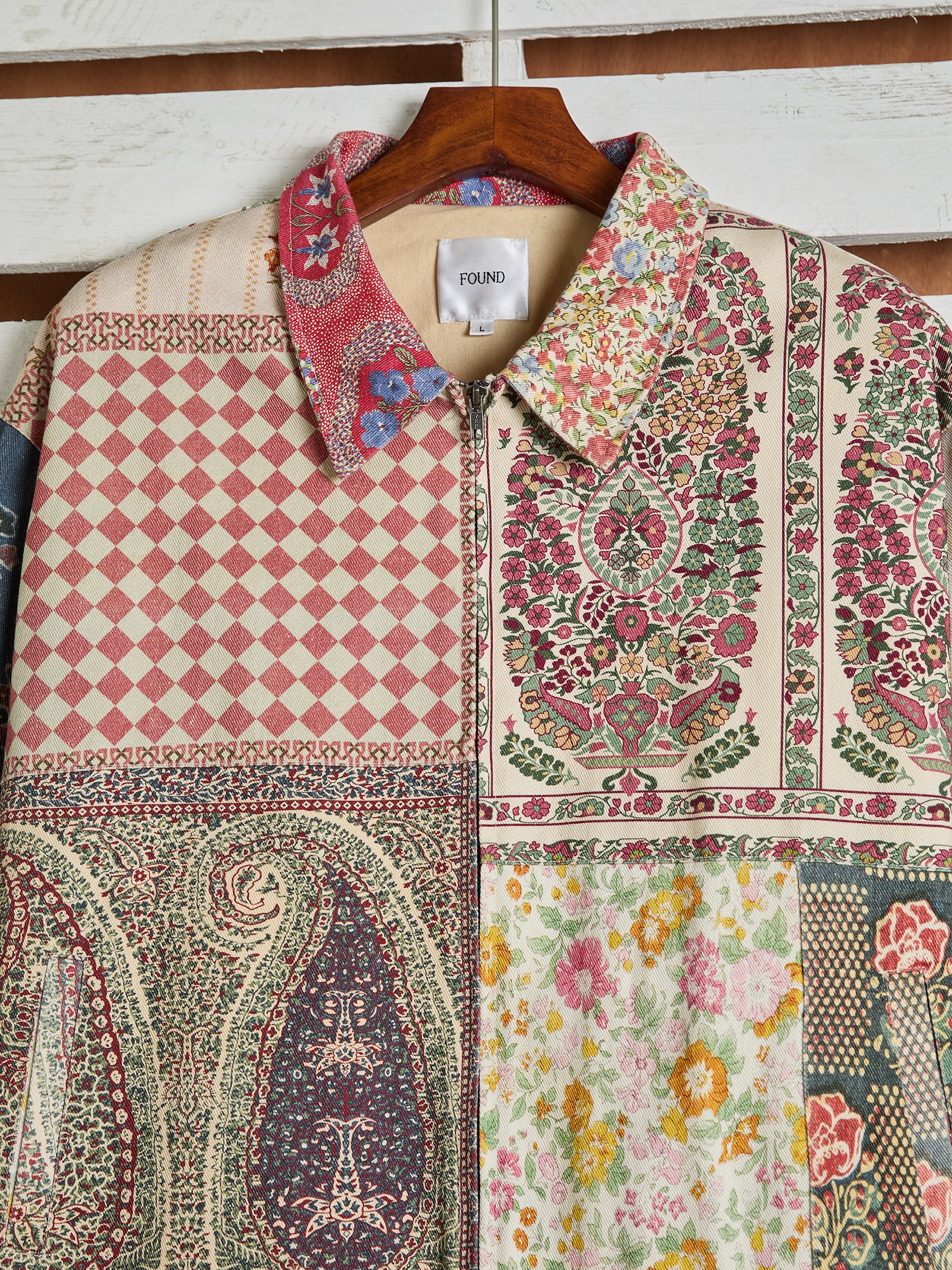 The Tapestry Twill Jacket by FOUND, featuring a variety of patterns like geometric, floral, and paisley prints for a vintage feel, hangs gracefully on a wooden hanger.