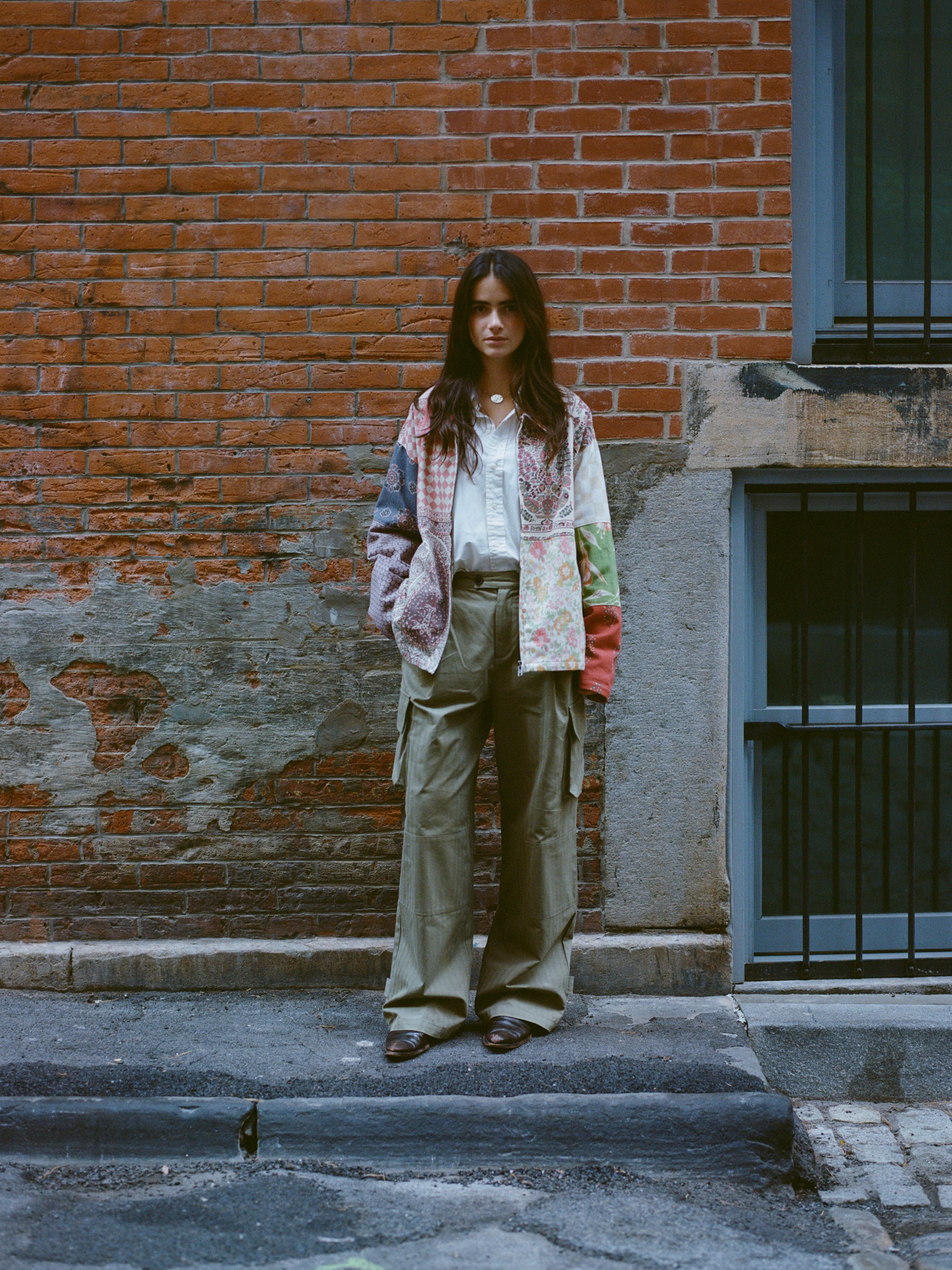 A person in front of a brick wall wears a vibrant FOUND Tapestry Twill Jacket, paired with a white top and cargo pants, merging modern flair with vintage charm.