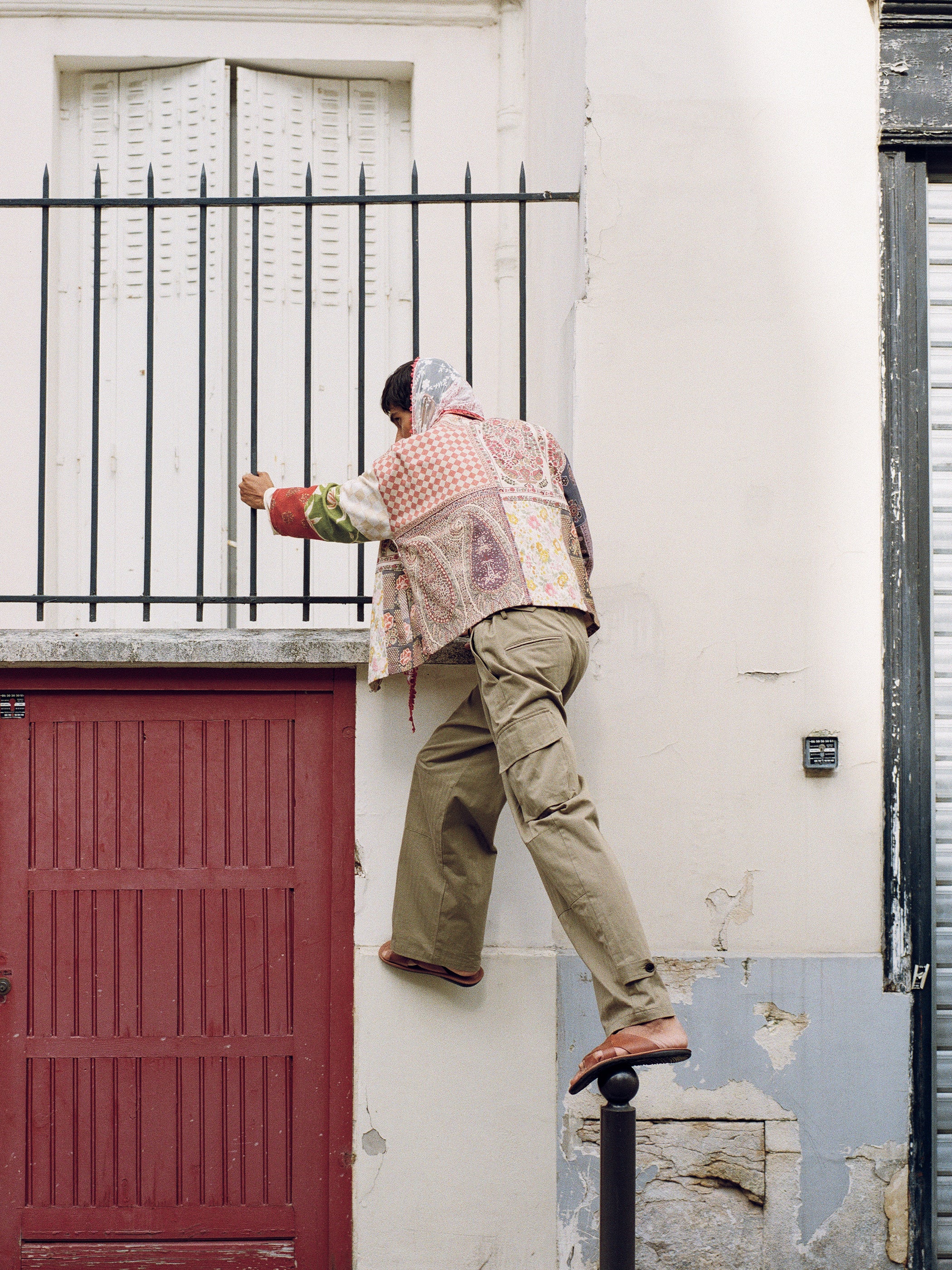 In an adventurous stride, a person climbs over a metal fence in the unisex relaxed fit of FOUND's Tapestry Twill Jacket with South Asian patterns, paired with khaki pants. One foot rests on a black post and the other on a red doorway ledge.