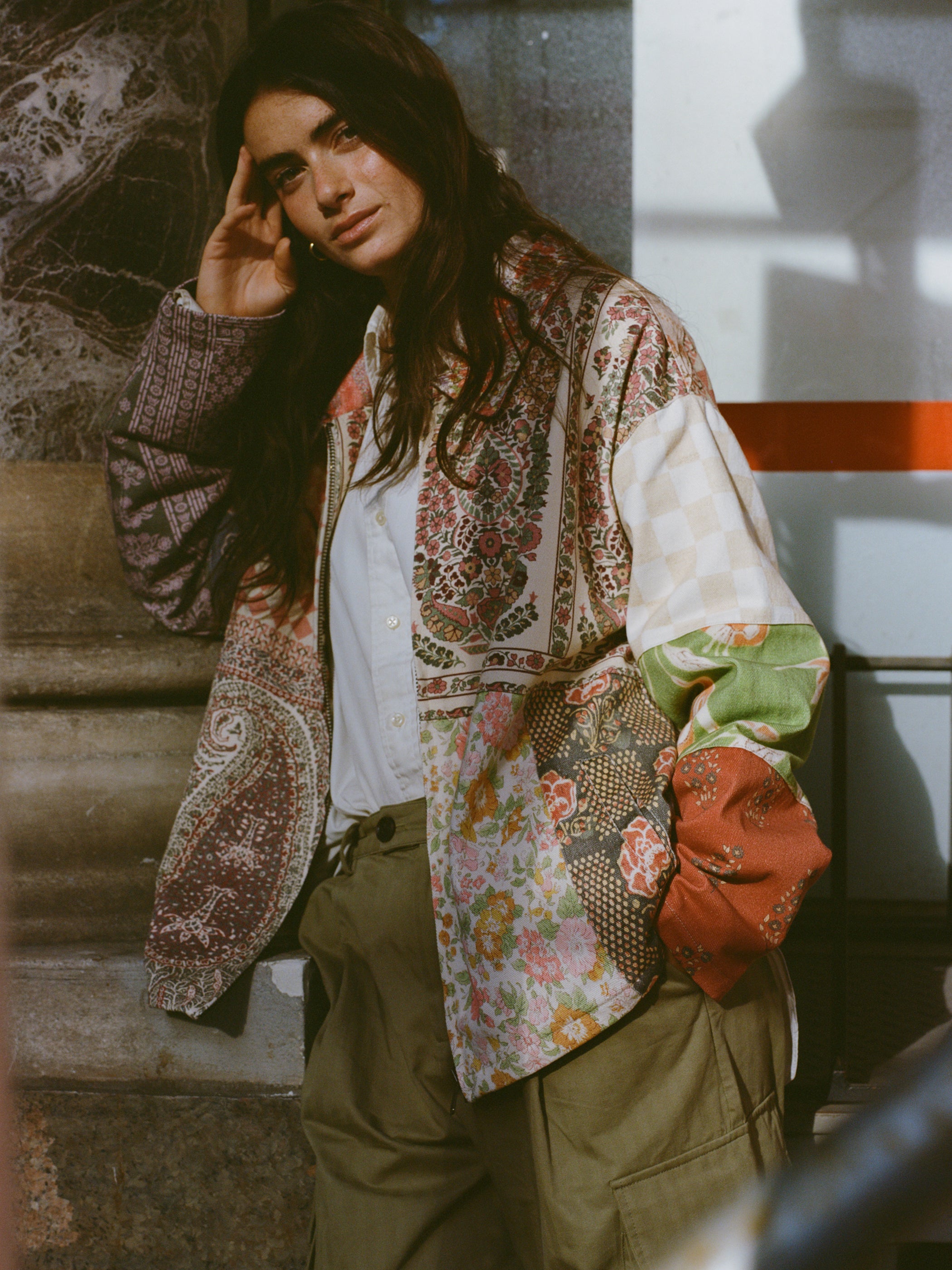 Wearing a vibrant Tapestry Twill Jacket by FOUND and khaki pants, a person leans thoughtfully against a marble structure with sunlight streaming in.