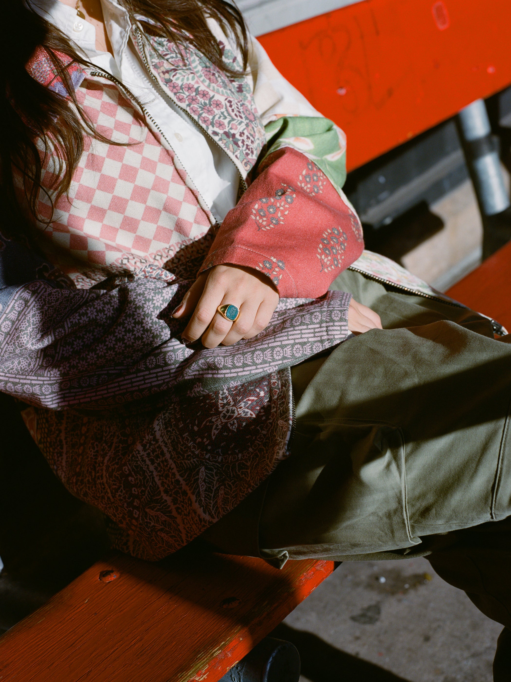 A person elegantly sits on a red bench, wearing a vintage Tapestry Twill Jacket by FOUND and a ring.