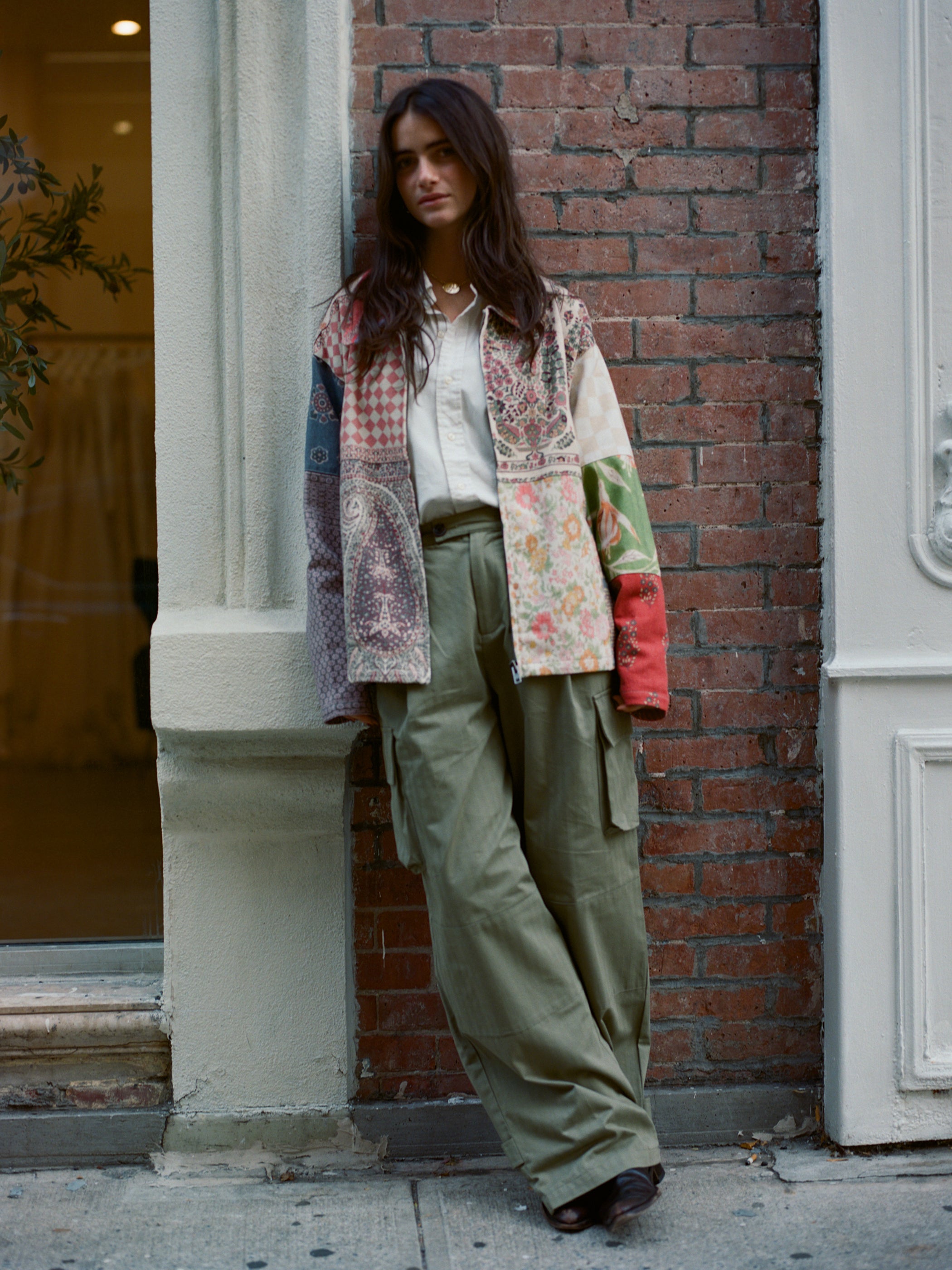 A woman in a FOUND Tapestry Twill Jacket and olive cargo pants leans against a brick wall on a city sidewalk, showcasing her timeless unisex style.