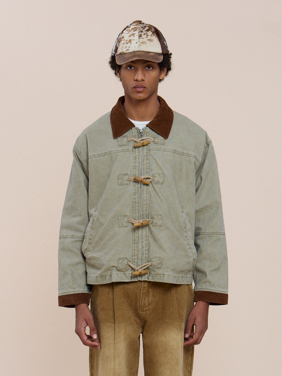 A person wears the FOUND Toggle Work Jacket in light denim with a brown collar and cuffs, tan pants, and a tan/brown cap against a beige background.