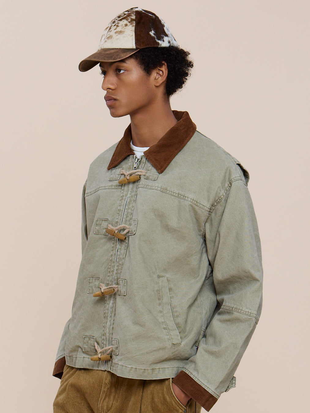 A person wears the FOUND Toggle Work Jacket in light green with a brown collar, tan pants, and a patterned cap, standing against a plain beige background.