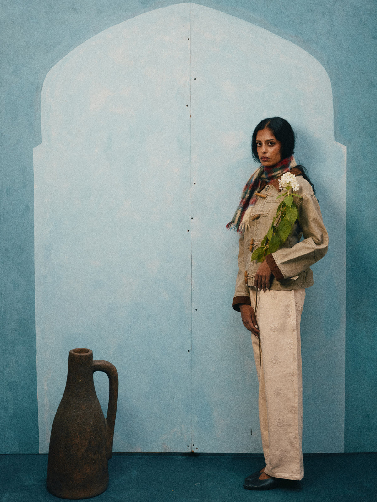 A woman in the FOUND Toggle Work Jacket stands holding flowers and a leafy branch before a blue arched wall, with a large brown jug on the floor beside her.