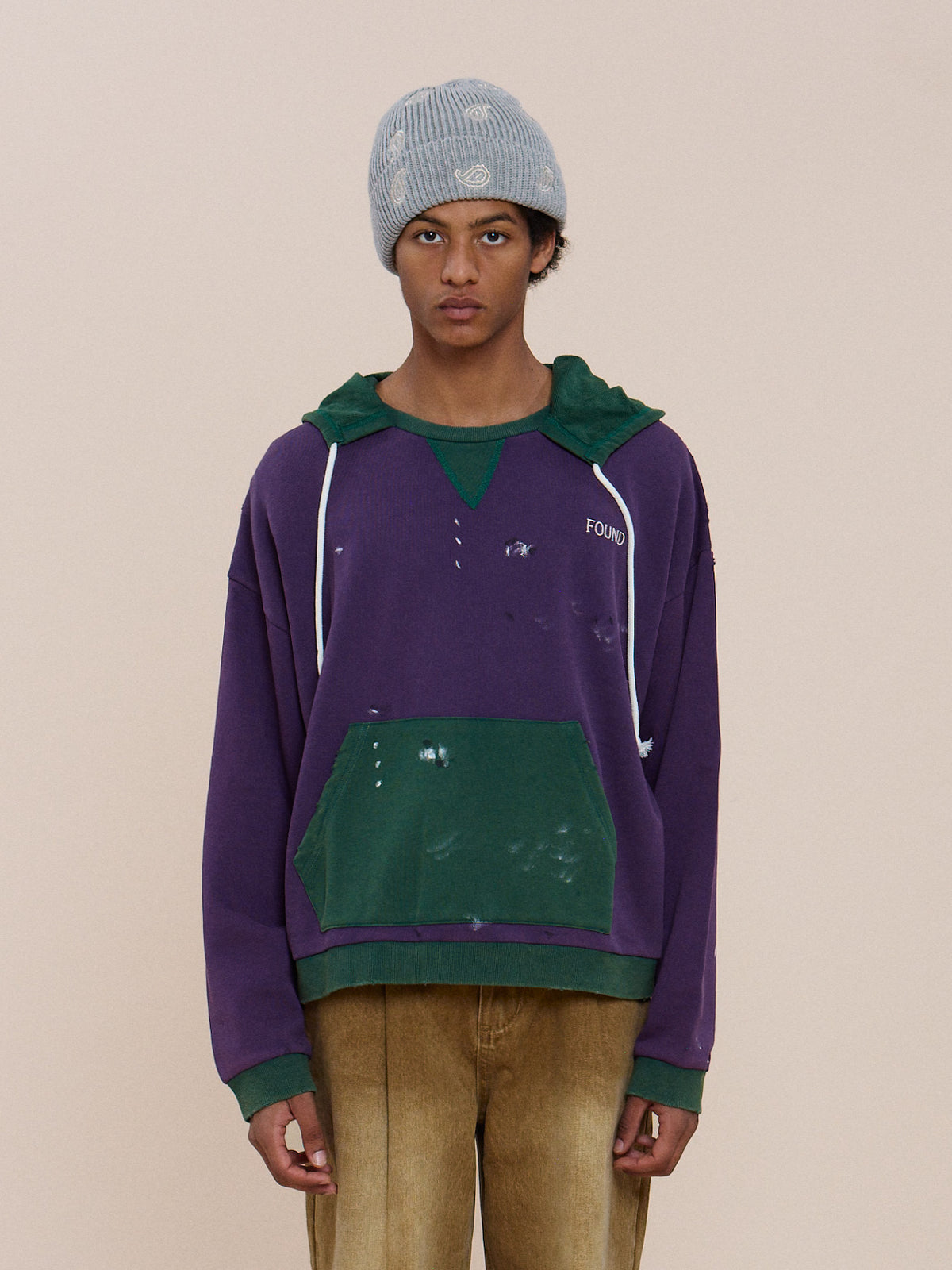 A person in a gray knit beanie and tan pants wears the FOUND Two Tone Painter Hoodie, featuring a unisex relaxed fit, against a plain beige background.