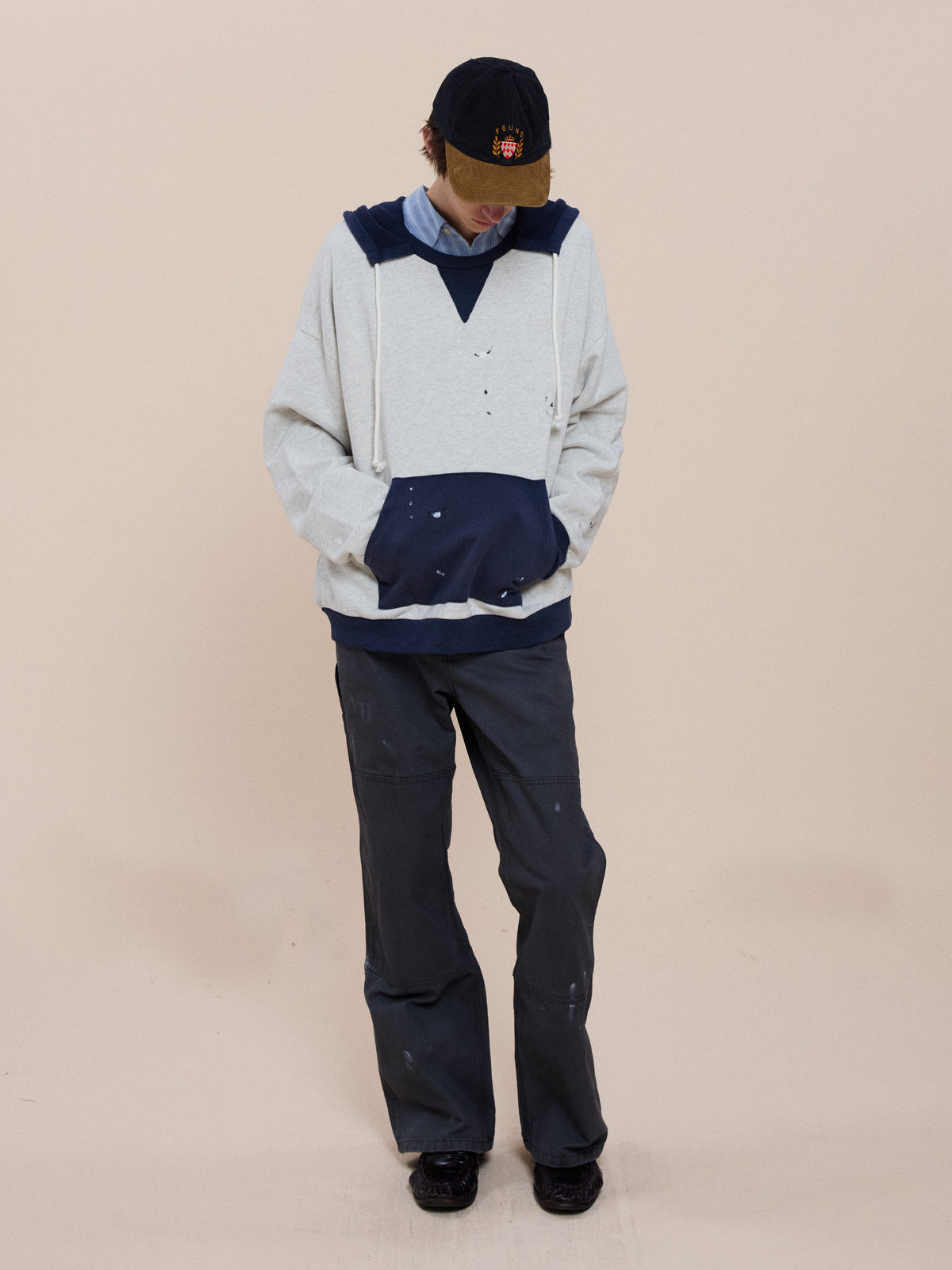 A person in a black cap, dark pants, and the FOUND Two-Tone Painter Hoodie (light gray/navy) stands with hands in the hoodie pocket against a beige background.