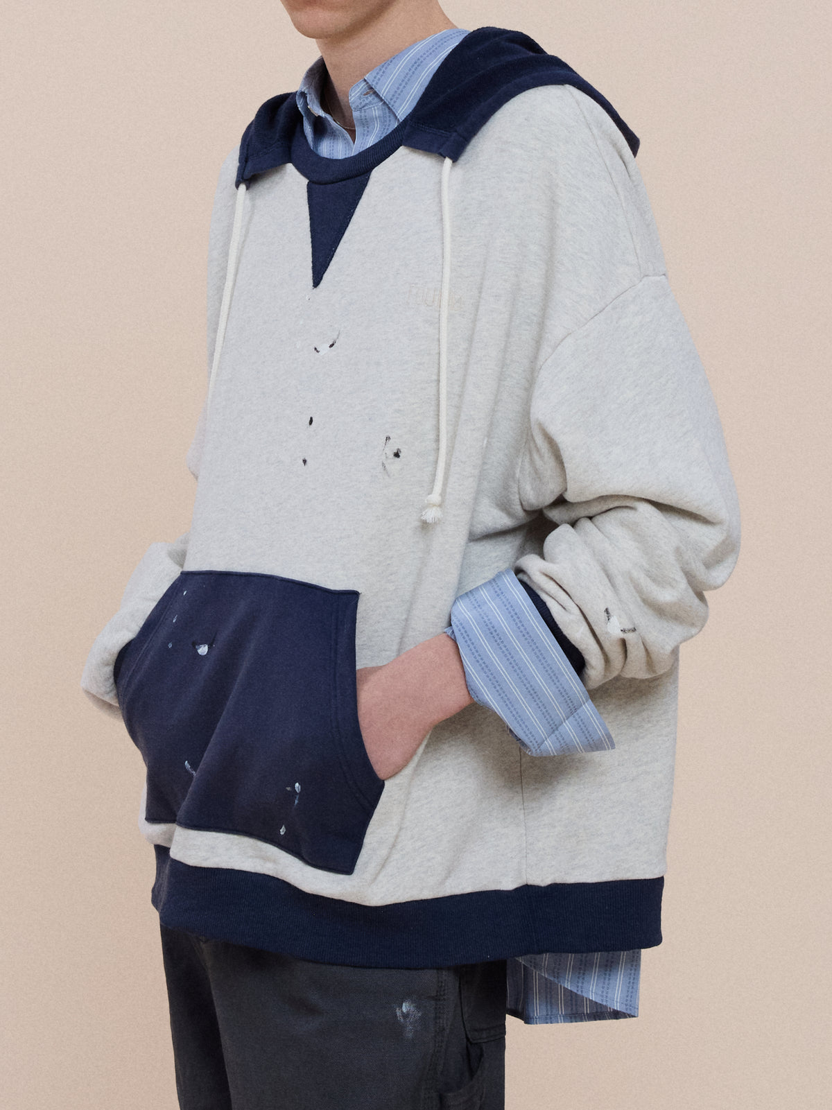 Wearing the FOUND Two-Tone Painter Hoodie—vintage grey with navy hood and pocket, relaxed fit, paint splatters, over a blue collared shirt.