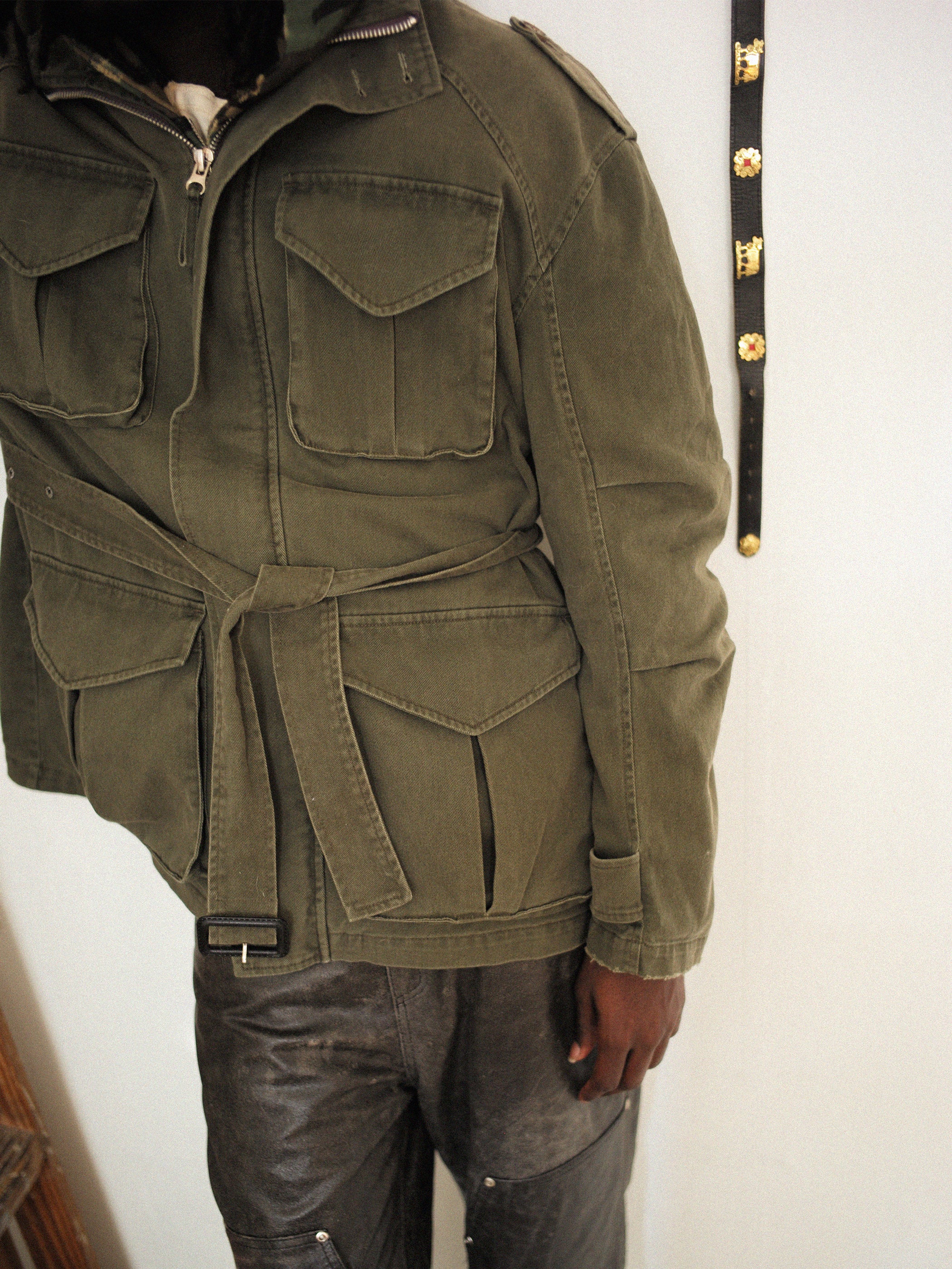 A person stands by a wall with a decorative belt hanging, wearing an M65 Utility Twill Pocket Jacket by FOUND and black pants, creating a vintage military-inspired look.