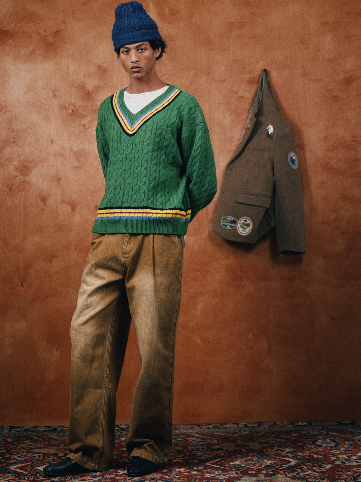 Person in a FOUND V-Neck Cricket Sweater, white shirt, loose tan pants, and blue beanie stands indoors; brown jacket with patches hangs on the wood-paneled wall behind them.