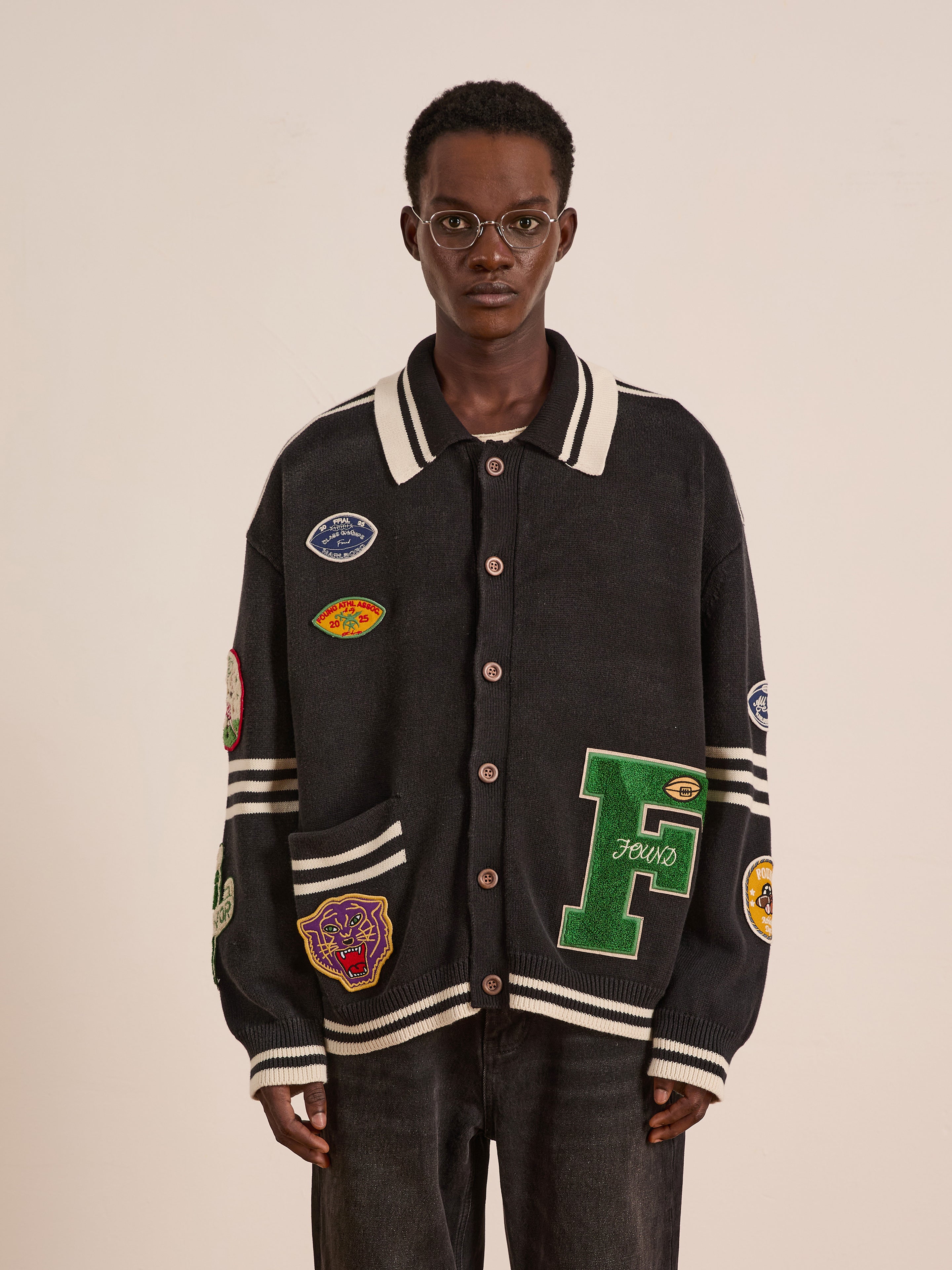 A person wearing glasses and FOUND's Varsity Patch Collared Cardigan, featuring assorted colorful patches, stands against a plain background.