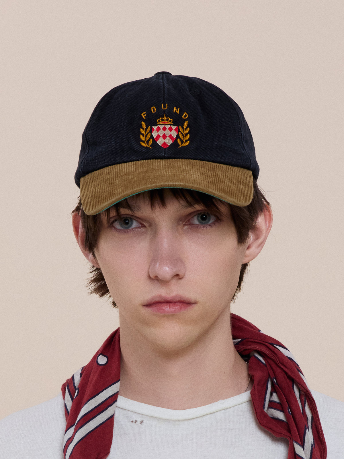 Person wears the Vintage Club Cap by Found, featuring vintage detailing and a “FOUND” crest, styled with a white shirt and red scarf against a neutral background.