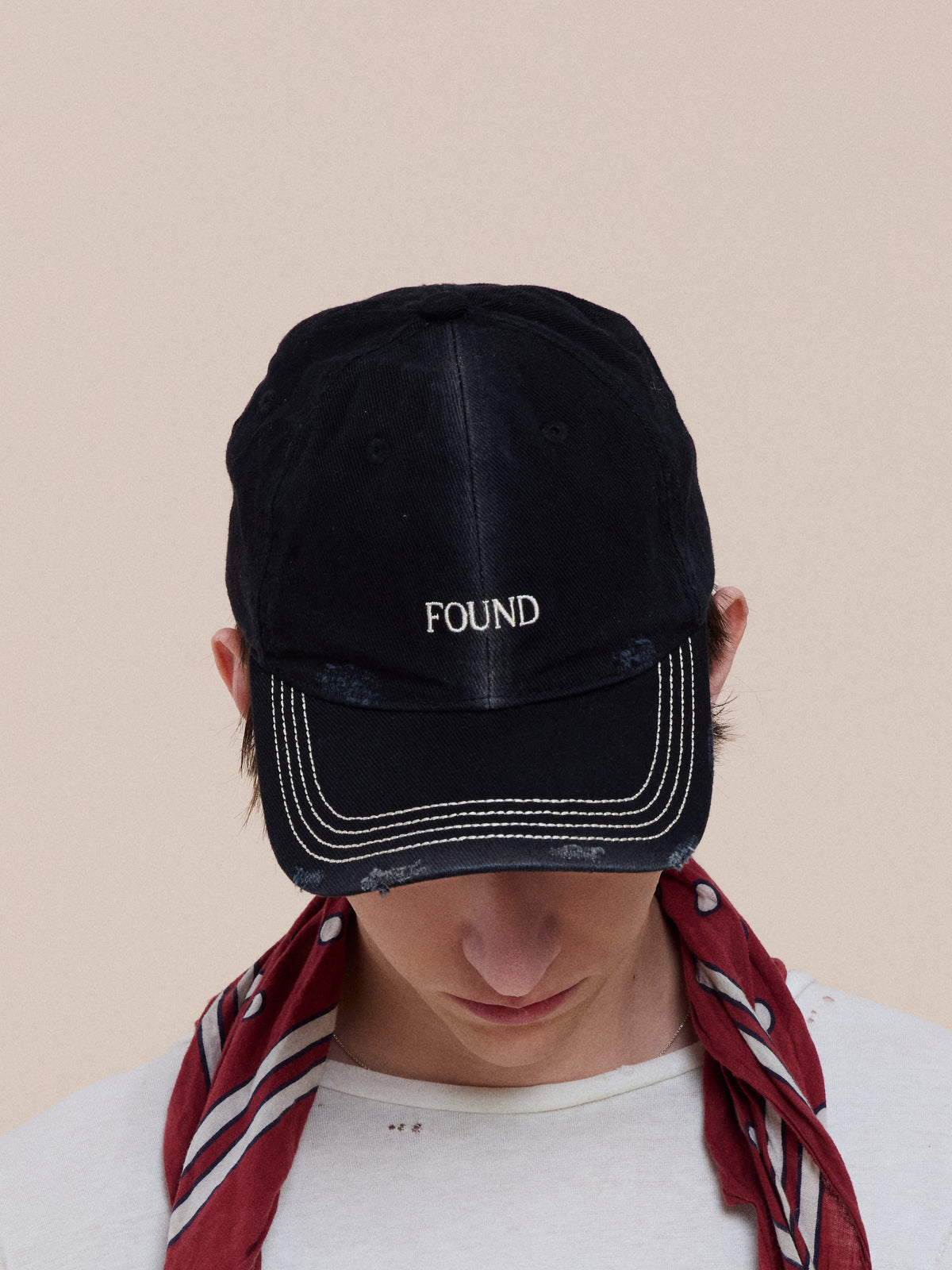 Wearing the Found Washed Logo Cap with “FOUND” embroidery, a person looks down; a red scarf and an adjustable leather strap complete the look.