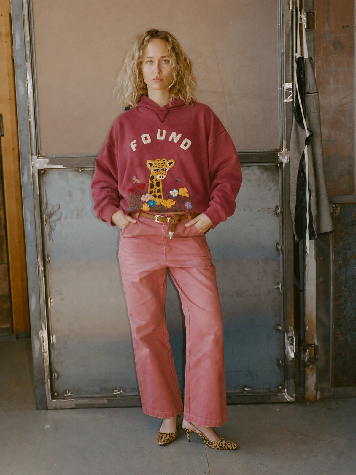 A person indoors wears a maroon FOUND Embroidered Wildlife Hoodie with a giraffe graphic, paired with pink pants and leopard print shoes, hands in pockets.