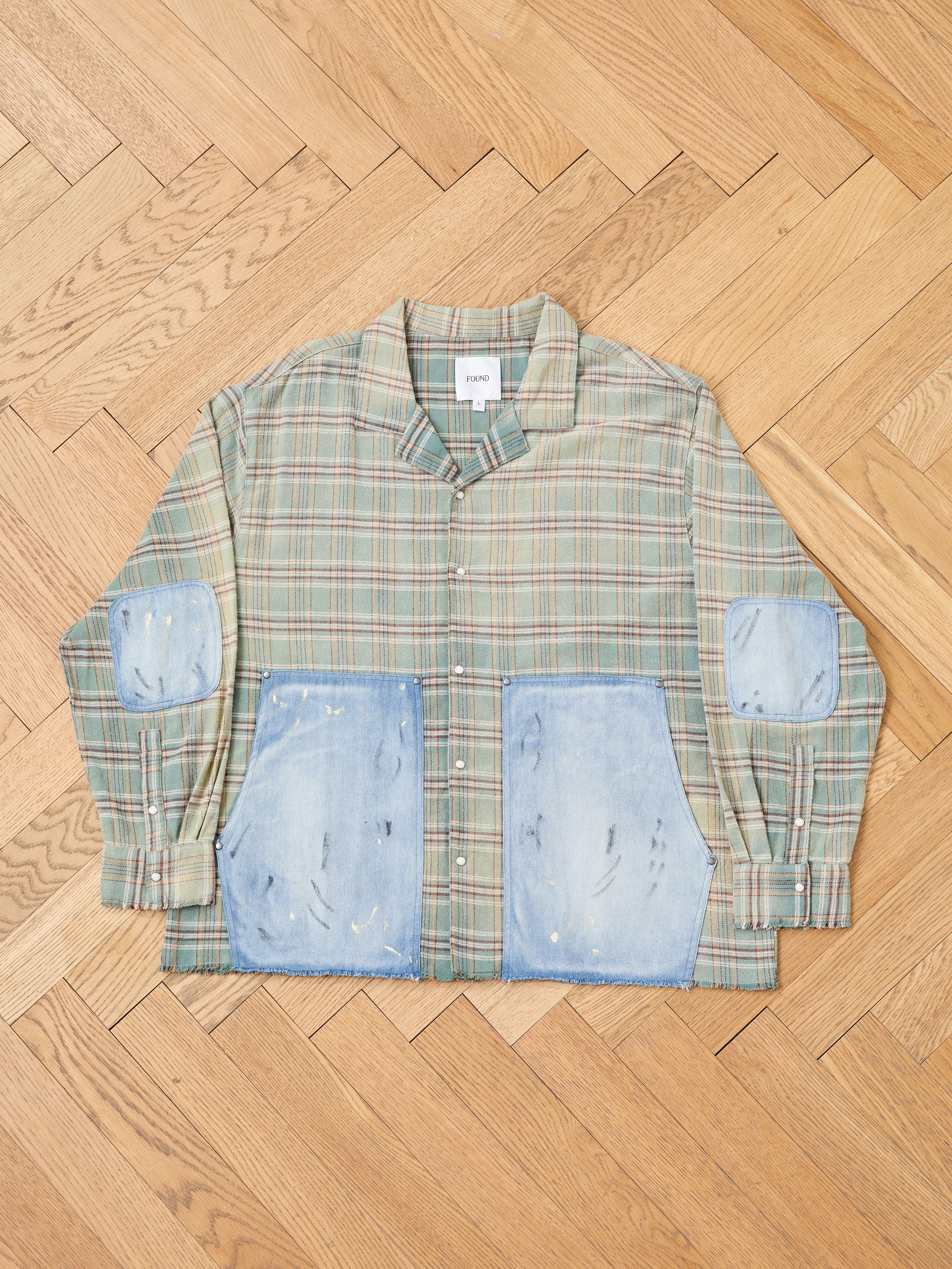 The FOUND Plaid Denim Shirt, a unisex green plaid shirt with denim panel pockets and elbow patches, is displayed flat on a parquet wooden floor.