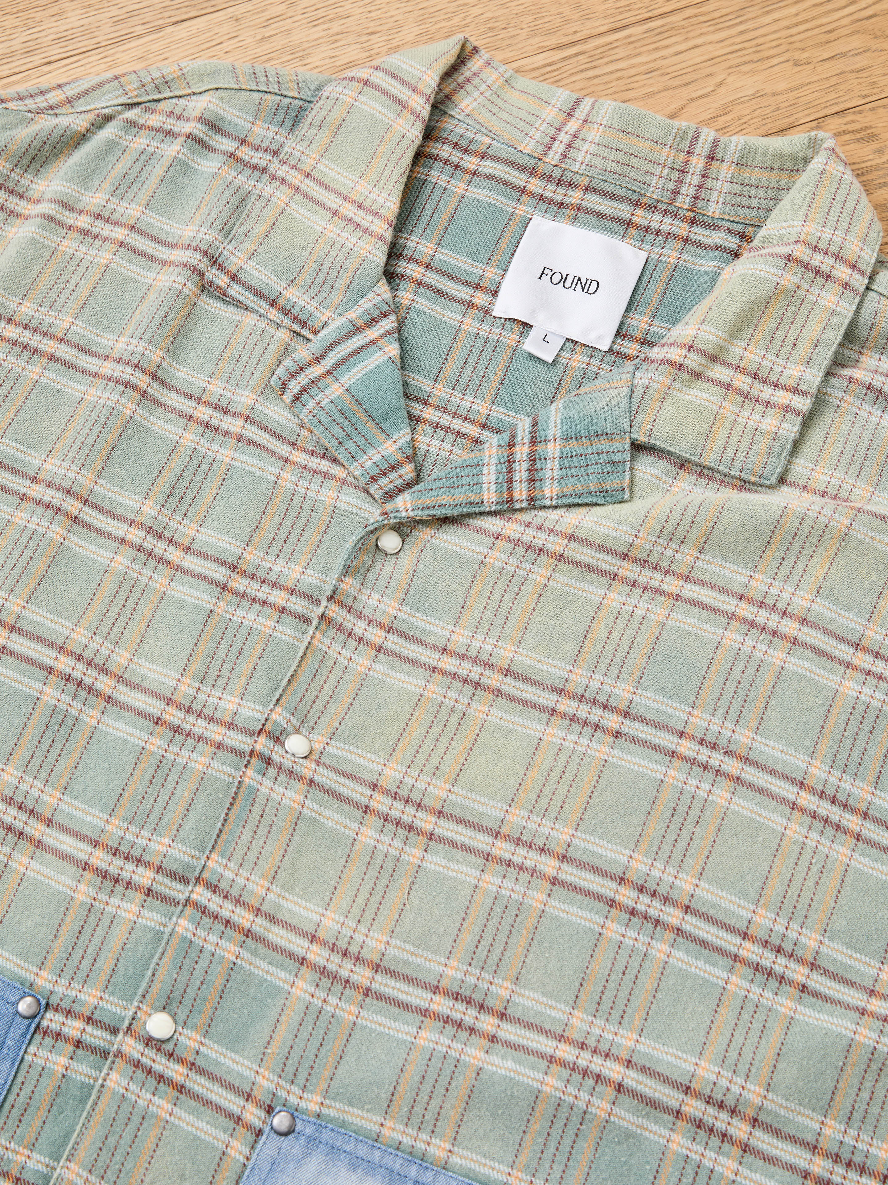 The FOUND Plaid Denim Shirt, with denim panel pockets and a "FOUND L" white label, is a stylish unisex workwear essential, shown laid flat on wood.