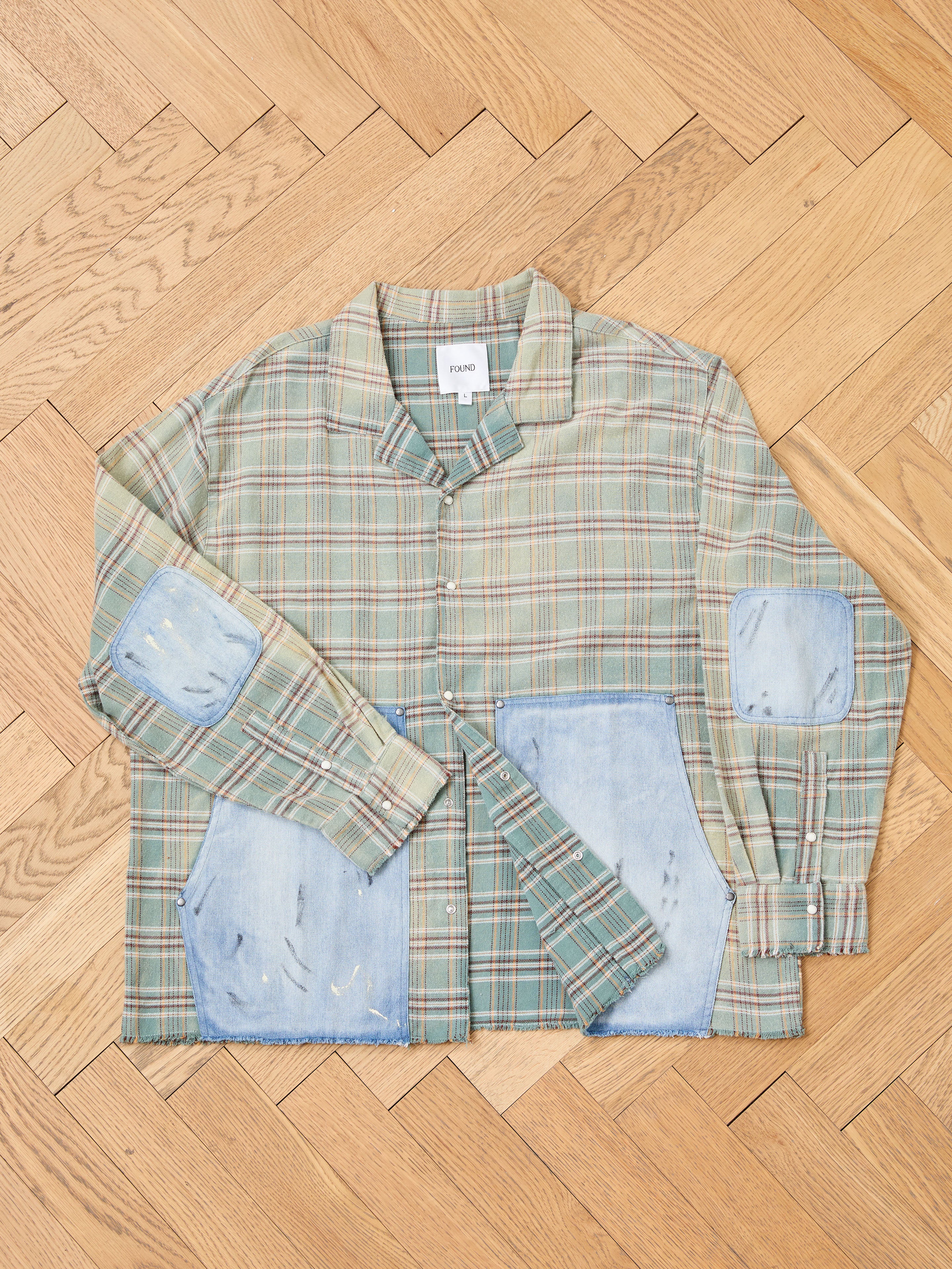 The FOUND Plaid Denim Shirt features green plaid, denim panel pockets, and ripped elbow patches, shown flat on a herringbone floor—a unisex workwear staple.