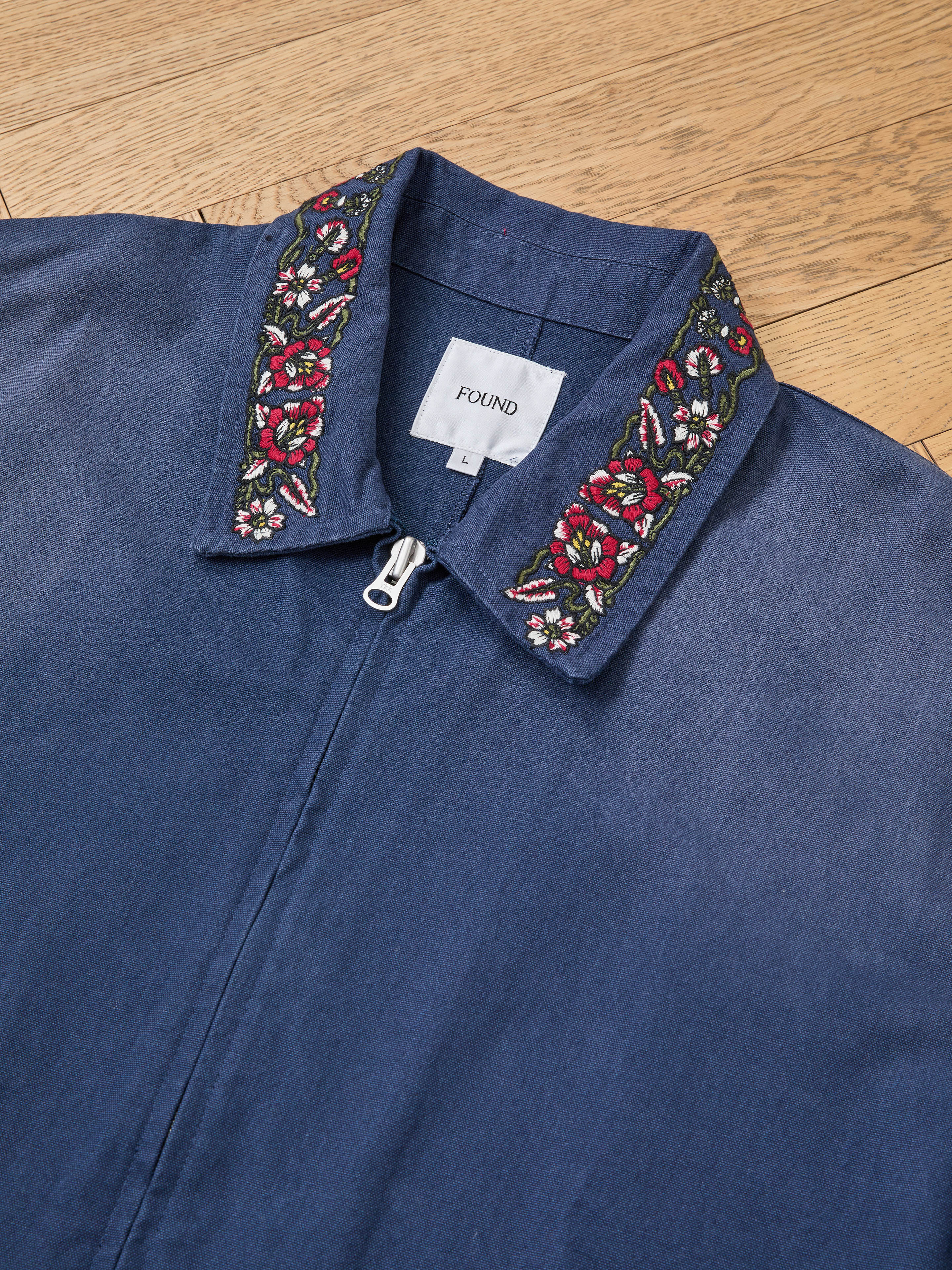 The Horse Equine Work Jacket by Found, a blue zip-up jacket with a floral embroidered collar, is displayed flat on a wooden surface. The inside label shows the brand name "Found.