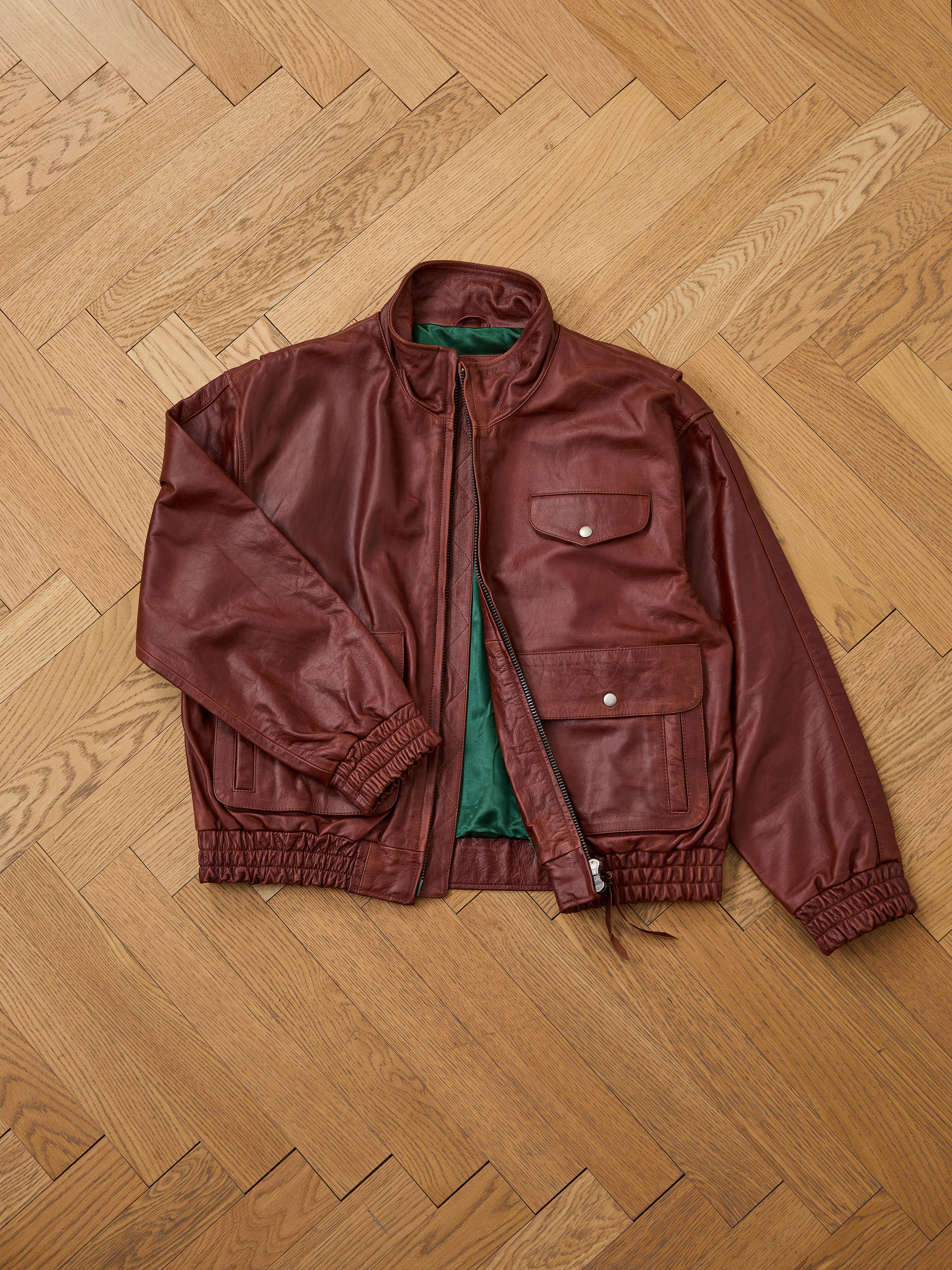 The Found Redwood Leather Bomber Jacket, a unisex brown leather piece with green lining, is laid flat on a light herringbone wooden floor. It features snap-button pockets plus elastic cuffs and hem for comfort and style.