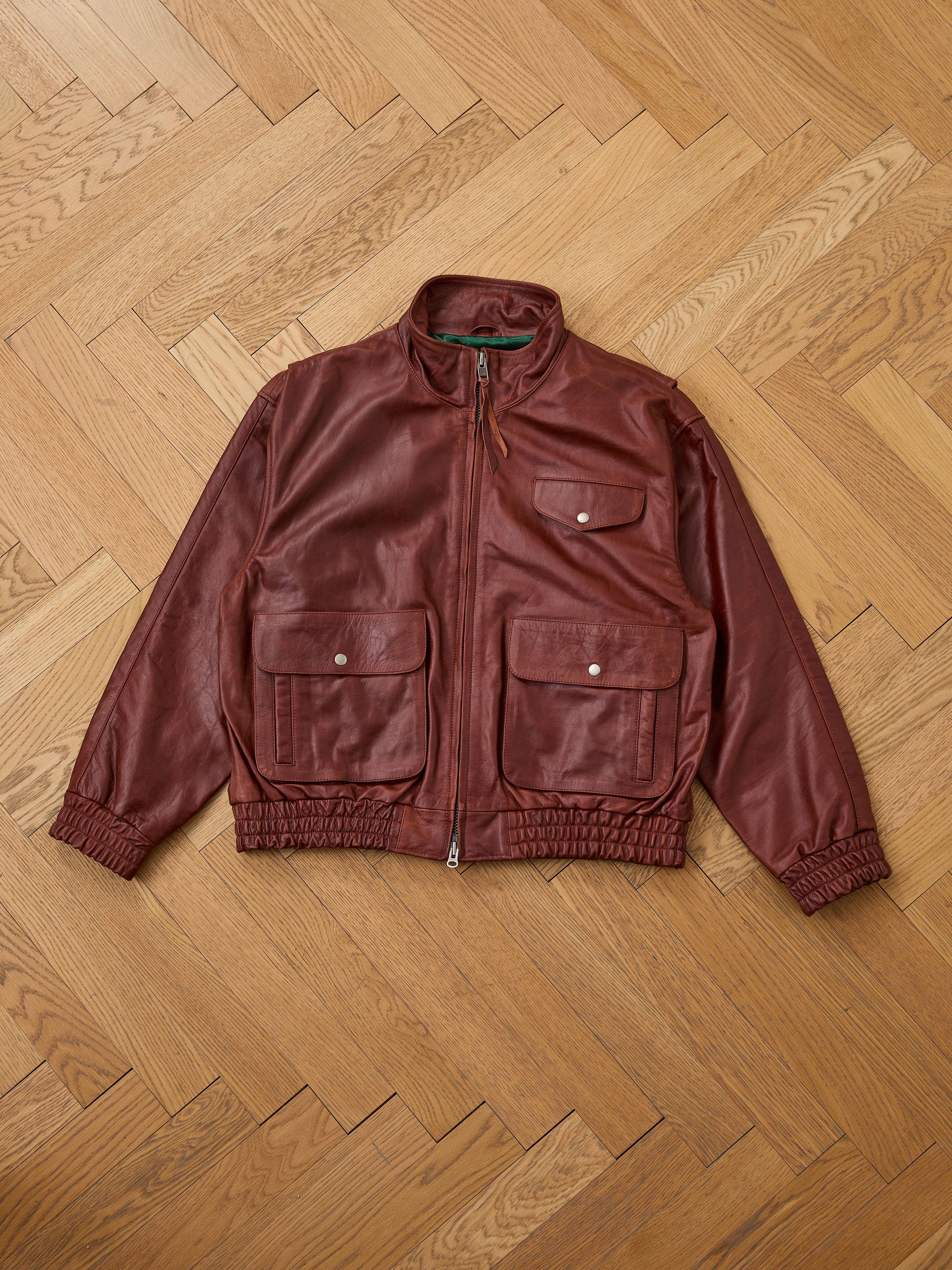 The Found Redwood Leather Bomber Jacket, shown laid flat on a light wood herringbone floor, features brown genuine leather, a front zipper, four flap pockets, and elastic cuffs for timeless unisex style.