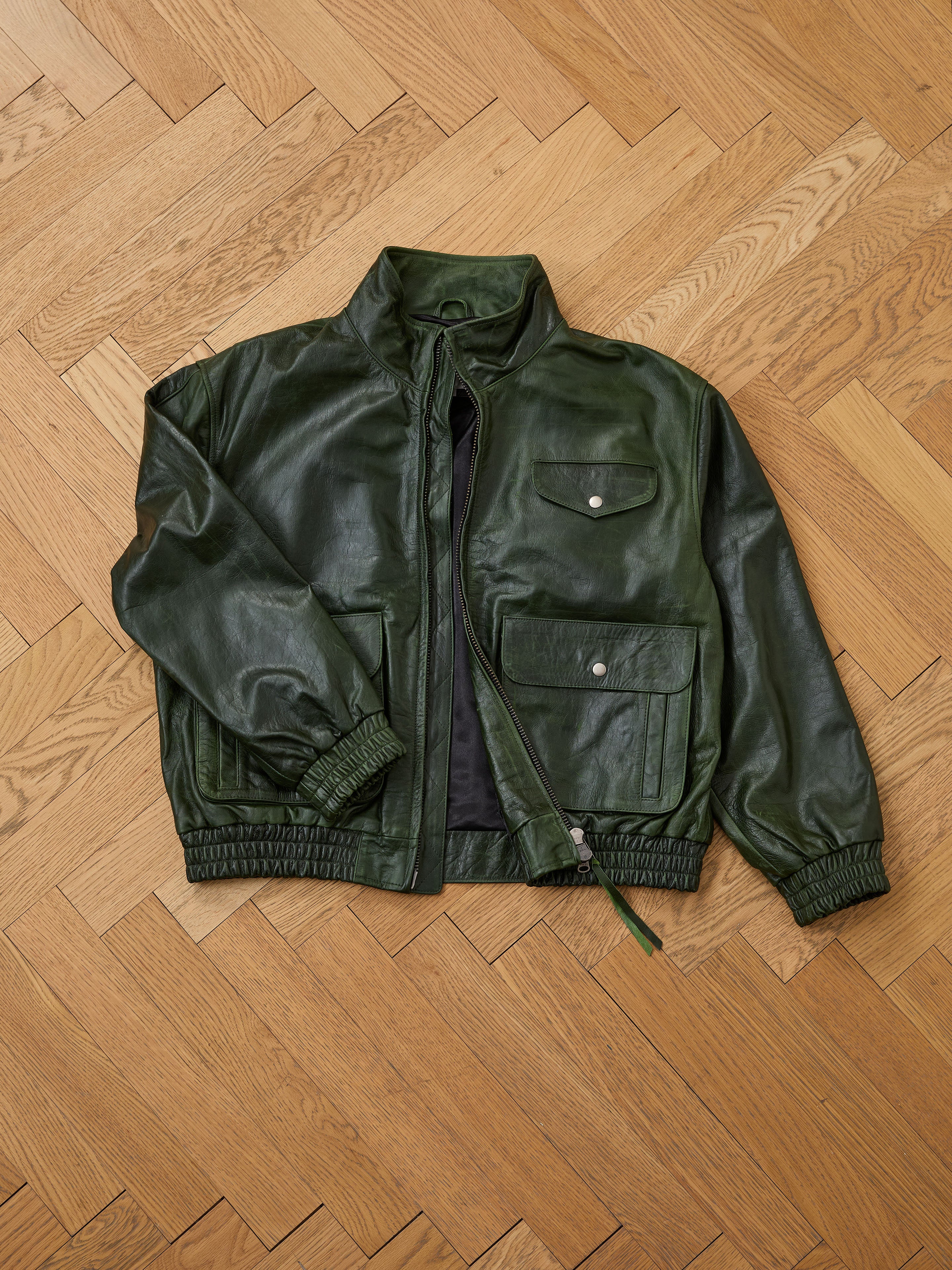 The FOUND Forest Leather Bomber Jacket, featuring snap button pockets and elastic cuffs, is laid flat on a wooden parquet floor.