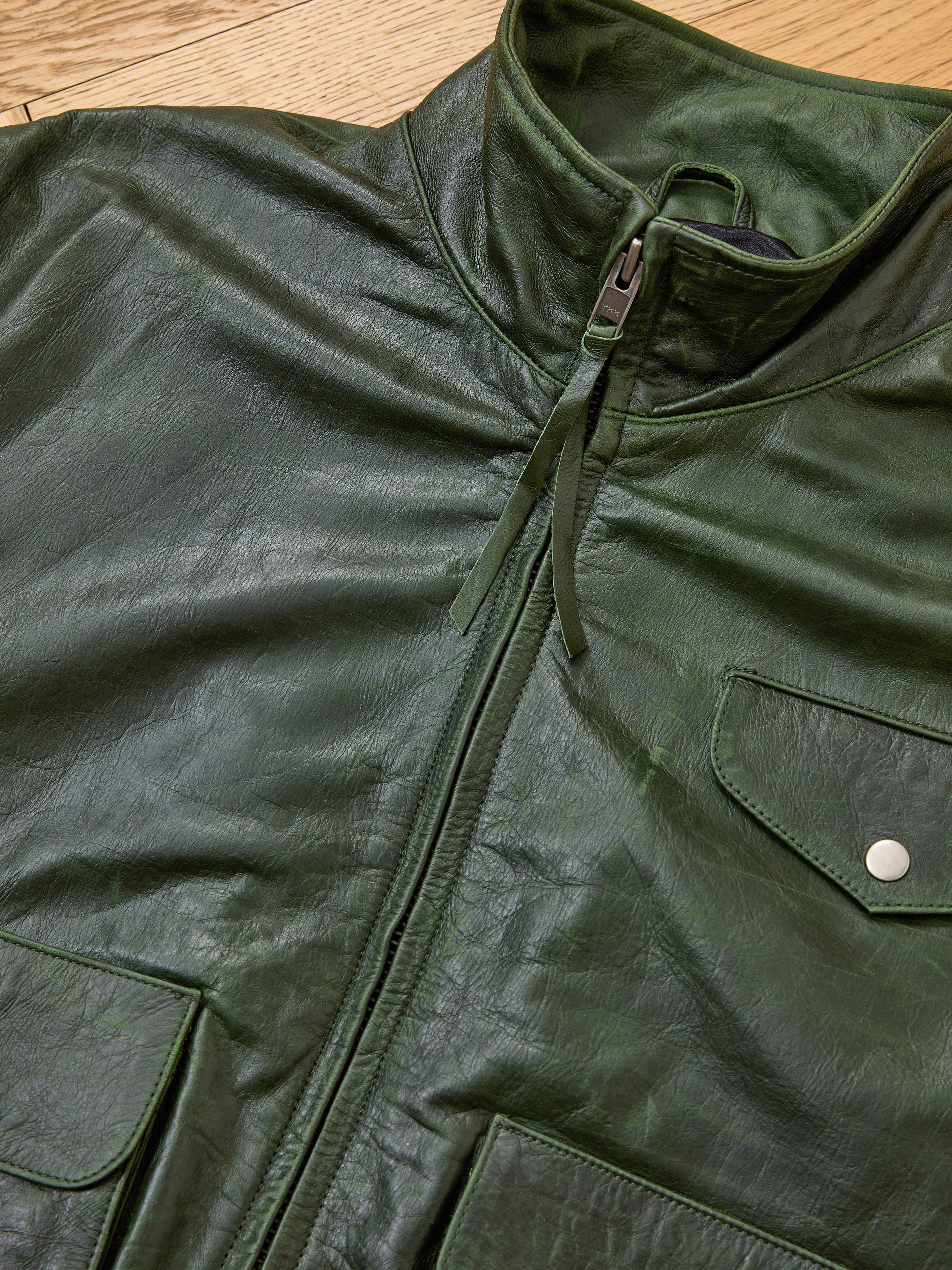 Close-up of the FOUND Forest Leather Bomber Jacket in dark green genuine leather, featuring a zippered front and snap-button flap pockets, laid flat on a wooden surface.