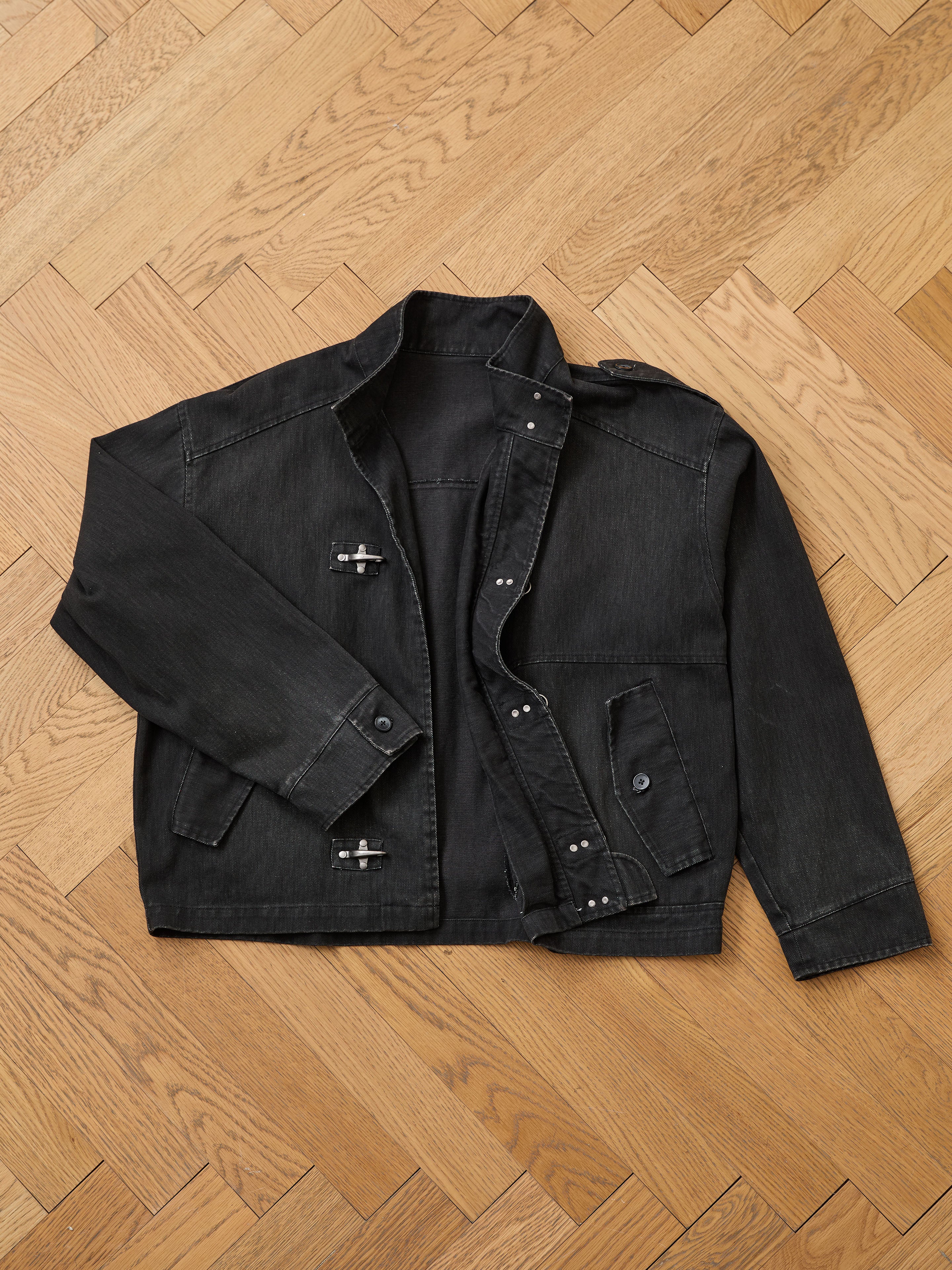 The Found Utility Clasp Work Jacket—a unisex, relaxed-fit black cotton twill piece with metal buttons and toggles—is displayed flat on a light herringbone wood floor.