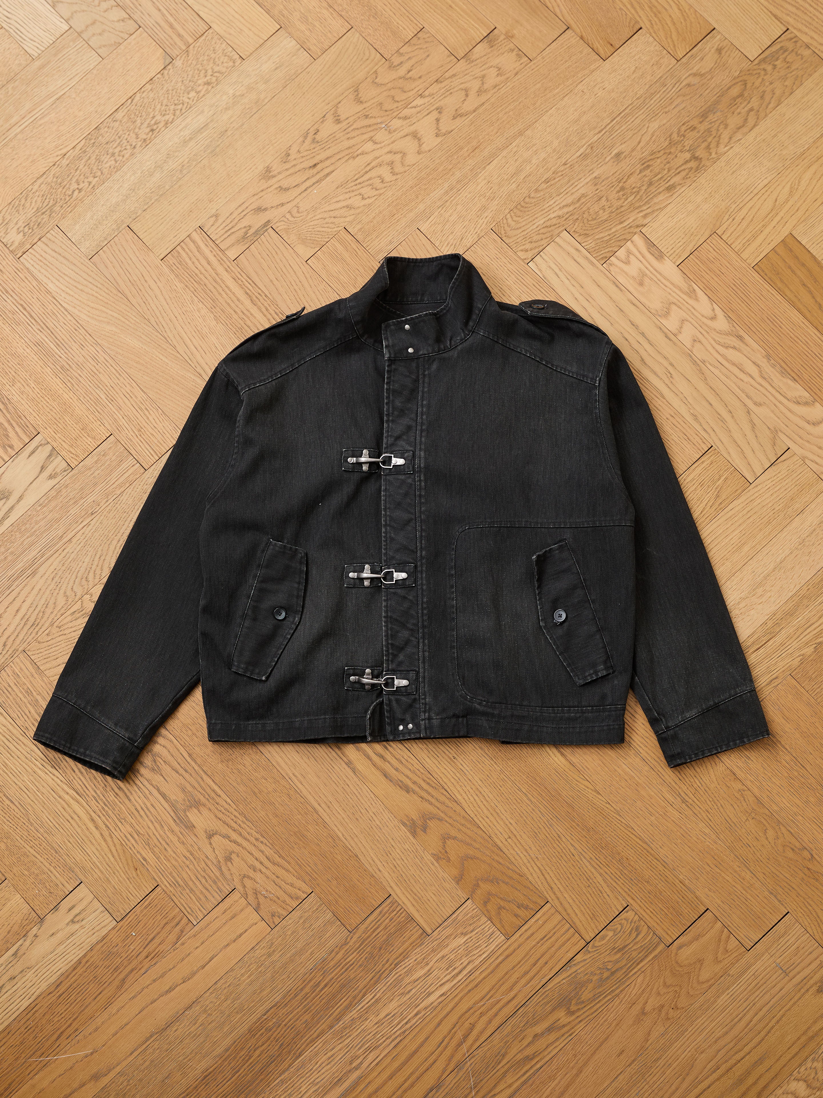 The Found Utility Clasp Work Jacket—a vintage-style, unisex relaxed fit in black cotton twill with silver toggles and two front pockets—is displayed flat on a wooden parquet floor.