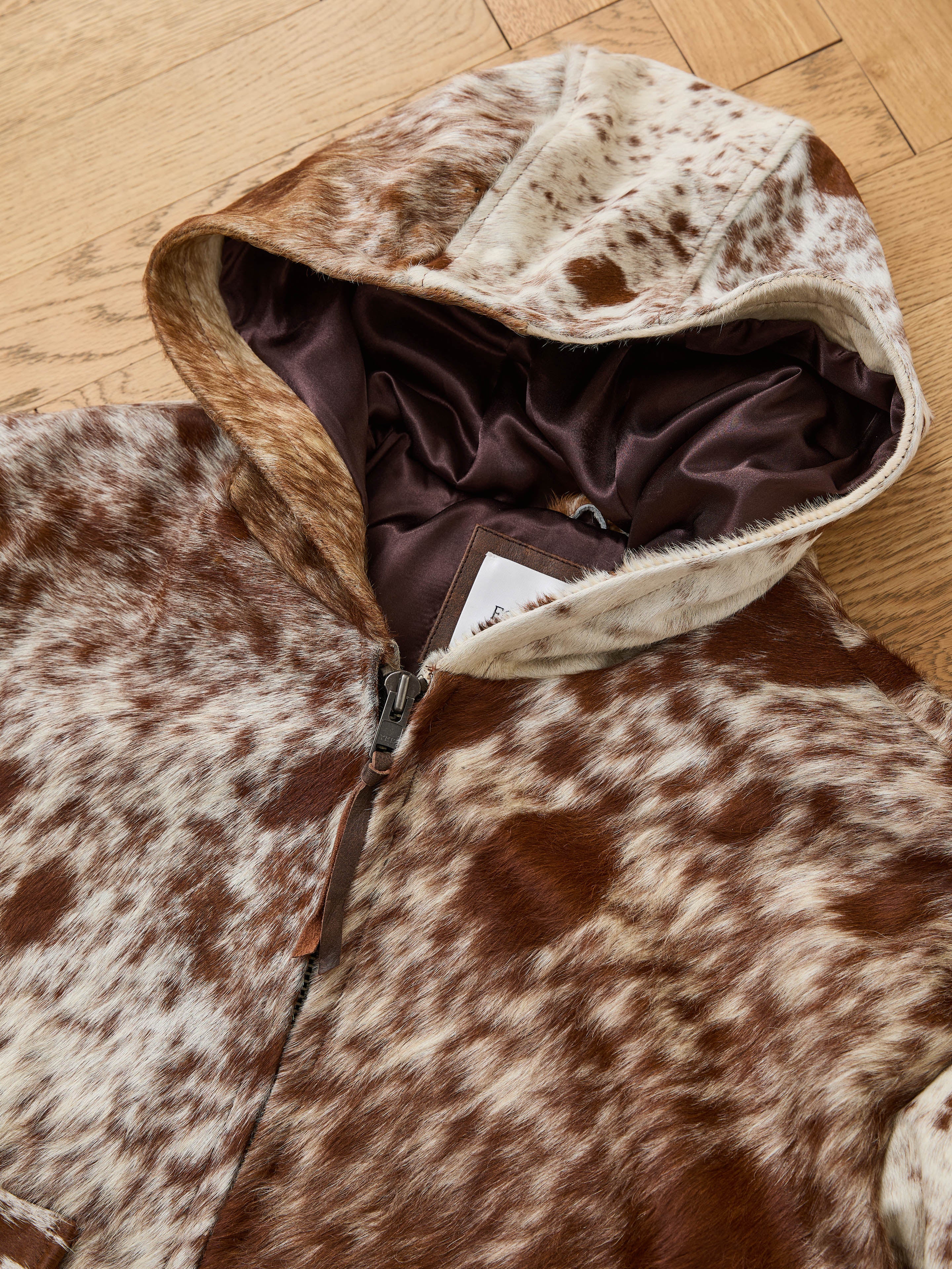The FOUND Cowhide Drover Hoodie features a brown and white spotted fur pattern and dark inner lining, shown partially unzipped and displayed on a wooden floor.