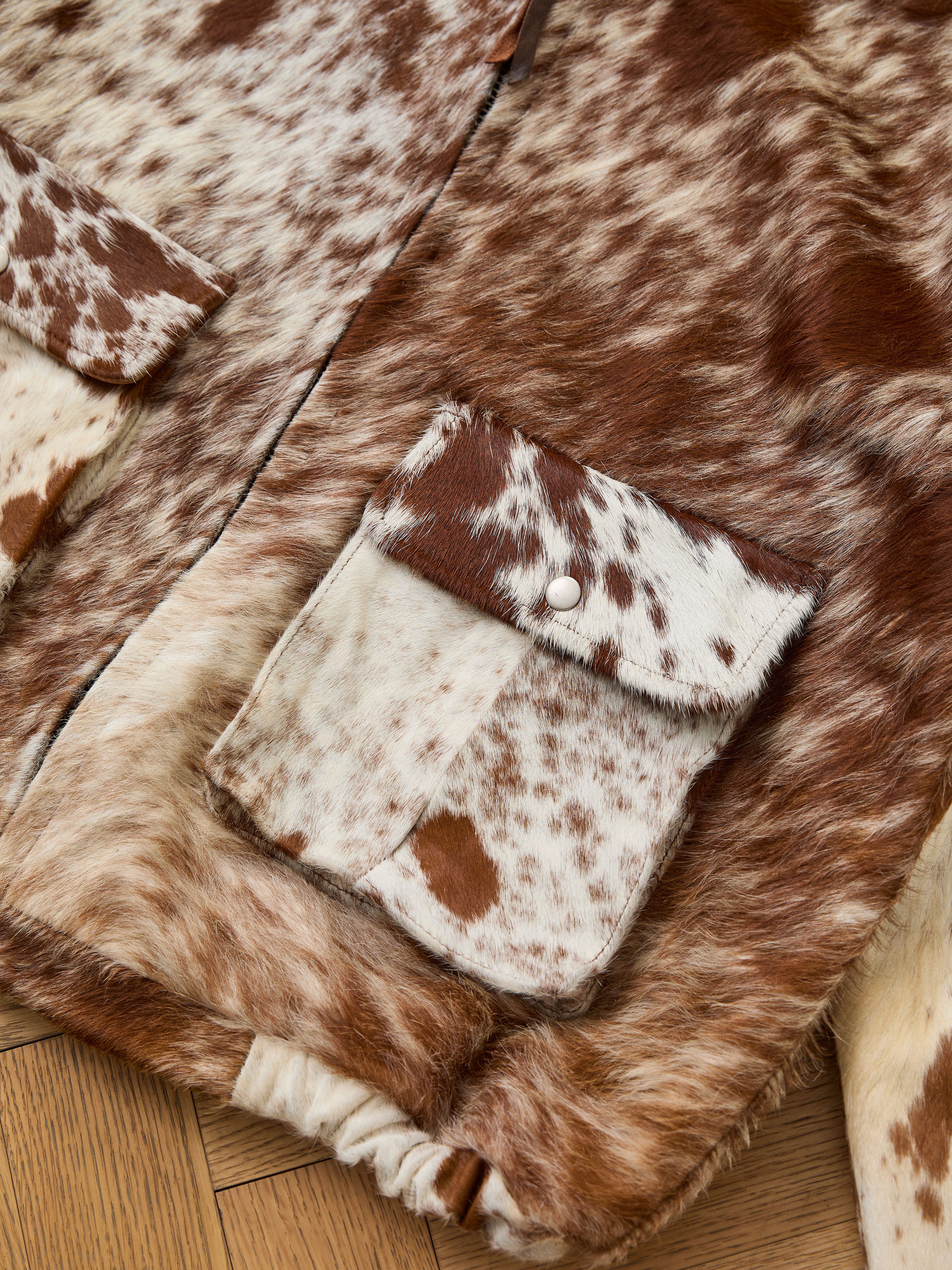 Close-up of the FOUND Cowhide Drover Hoodie, a brown and white faux fur jacket inspired by genuine cowhide, featuring a buttoned flap pocket, displayed on a wooden floor.