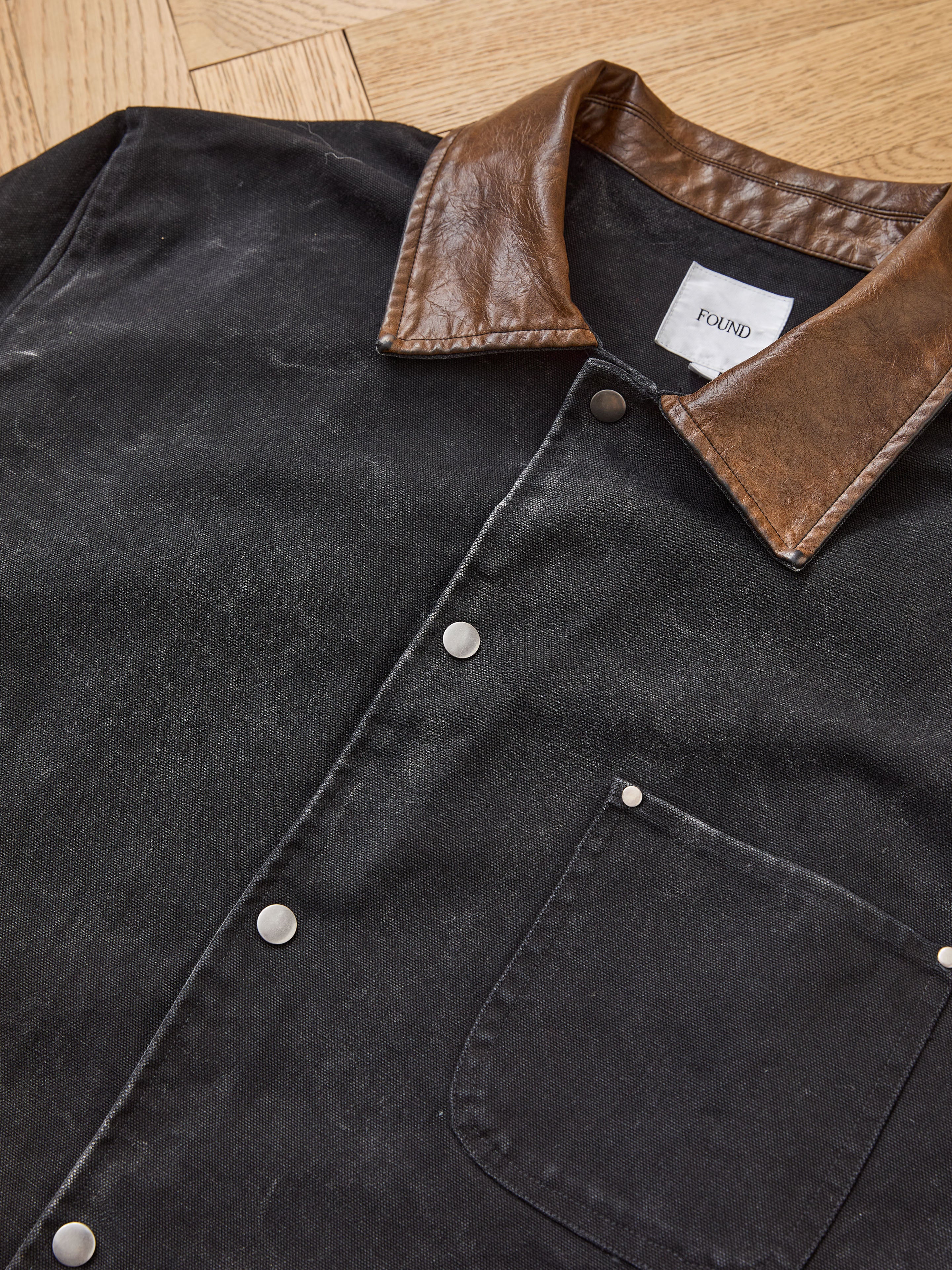 The Found Canvas Embroidered Work Jacket, featuring a brown leather collar, metal snap buttons, leather trim, and a chest pocket, is displayed flat on a wooden surface.