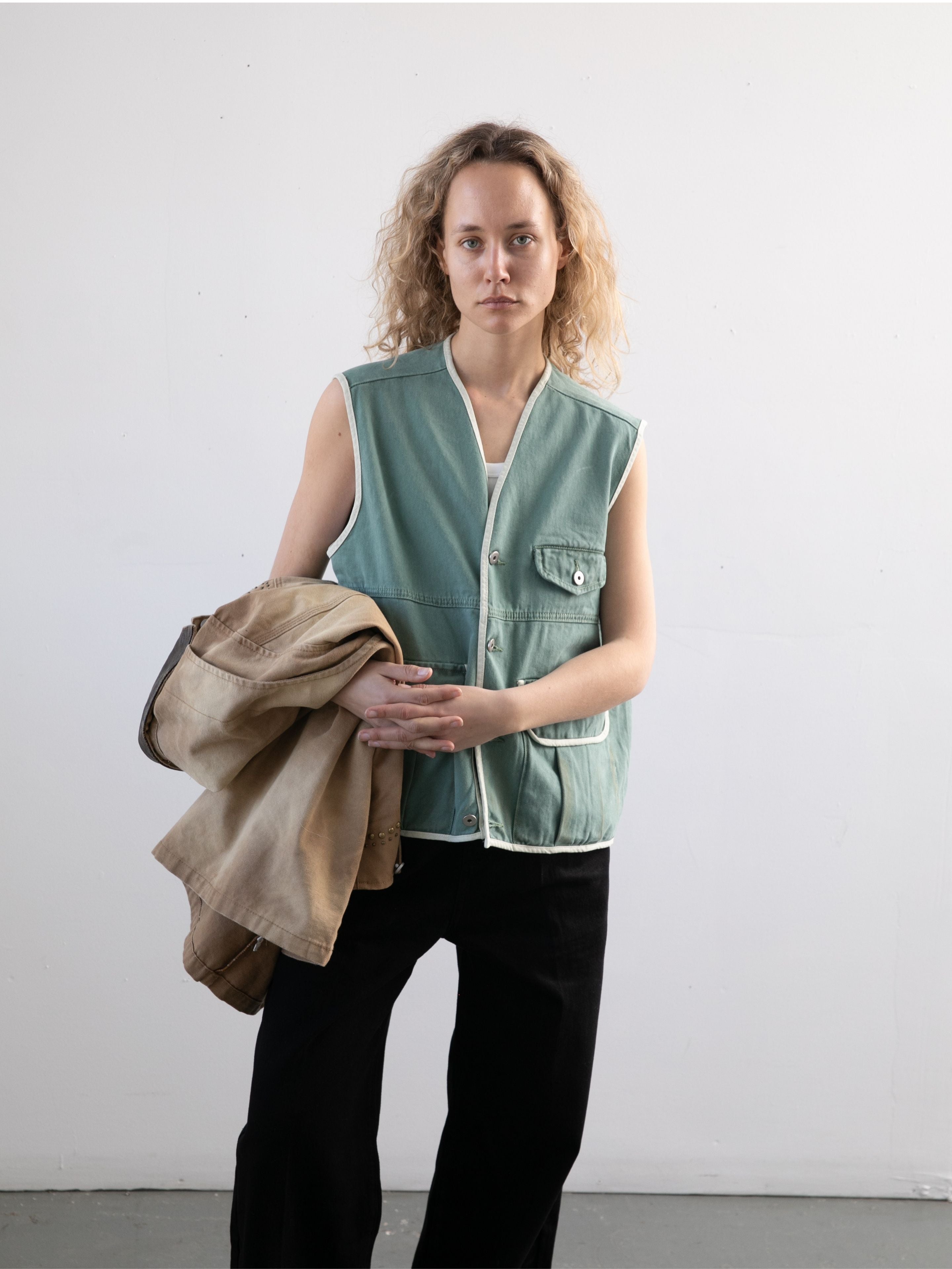 A person with curly hair stands against a plain wall, wearing the FOUND Cotton Twill Utility Vest in vintage washed green and black pants, holding a tan jacket in one arm.