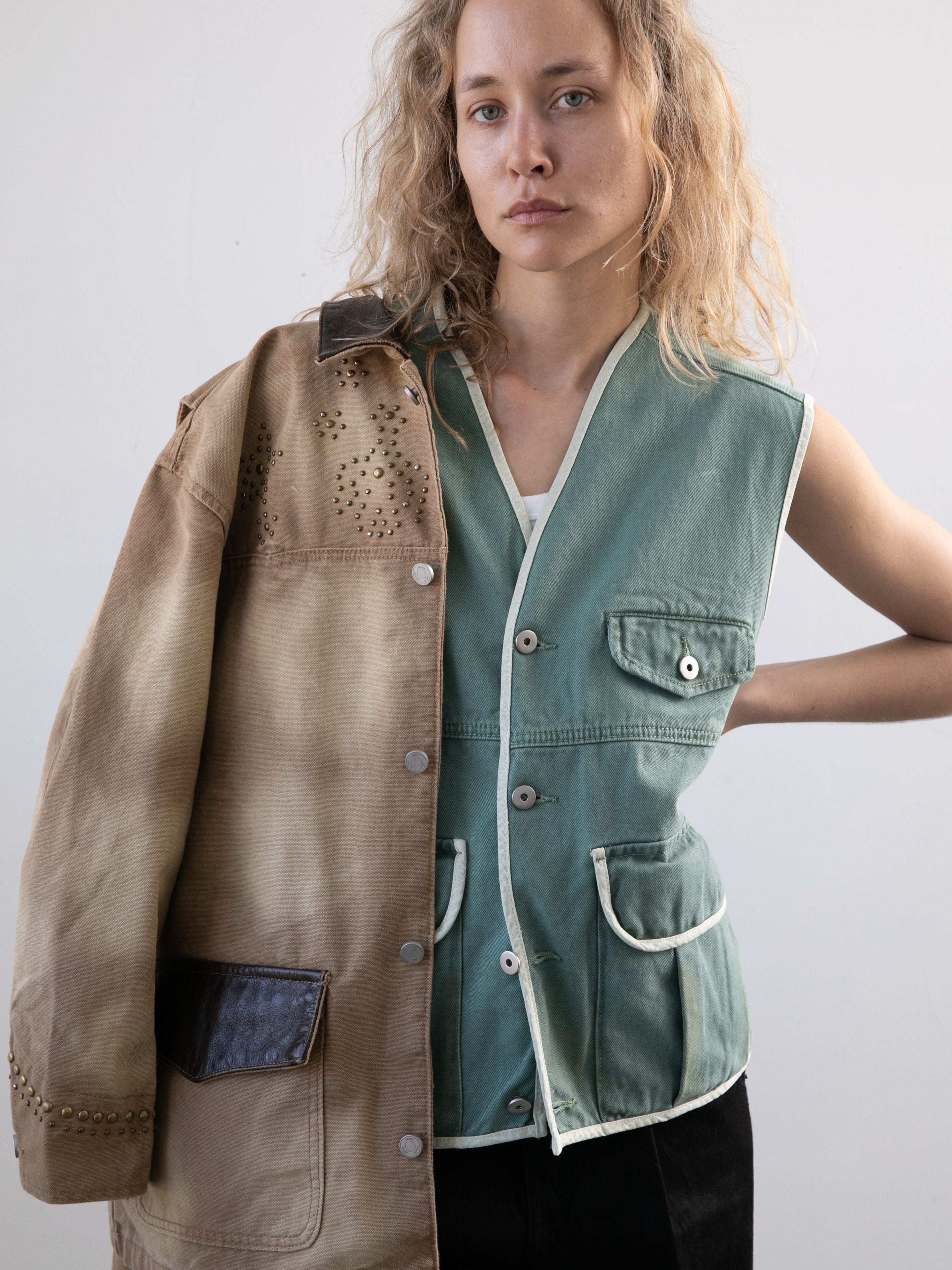 A person with wavy blonde hair wears the FOUND Cotton Twill Utility Vest in vintage washed green over a white shirt, draping a tan studded jacket over one shoulder, standing against a plain background.