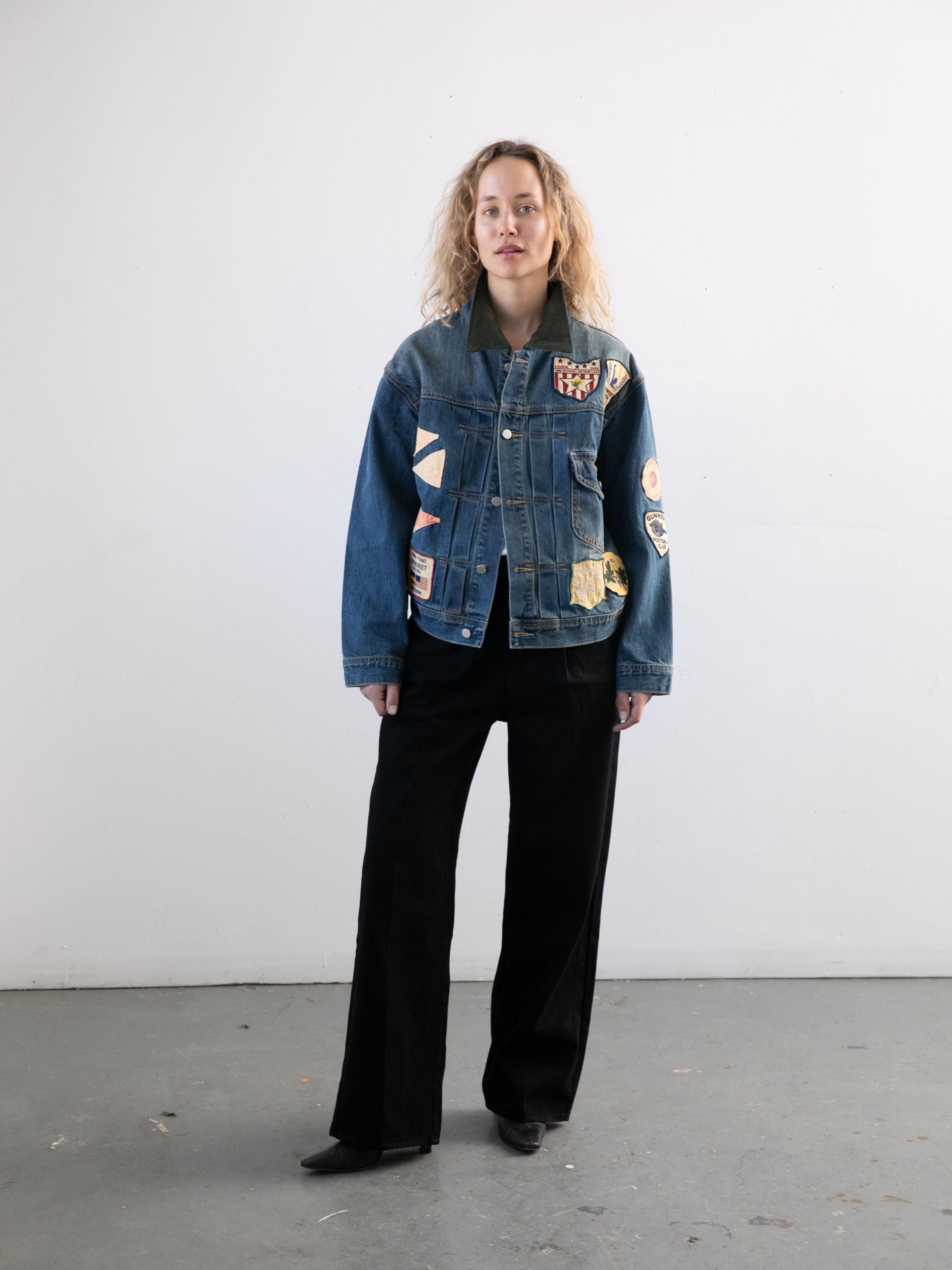 A person stands against a plain wall wearing the FOUND Denim Patchwork Jacket, styled in unisex relaxed fit with vintage wash and patches, paired with black pants and boots for a classic workwear-inspired look.