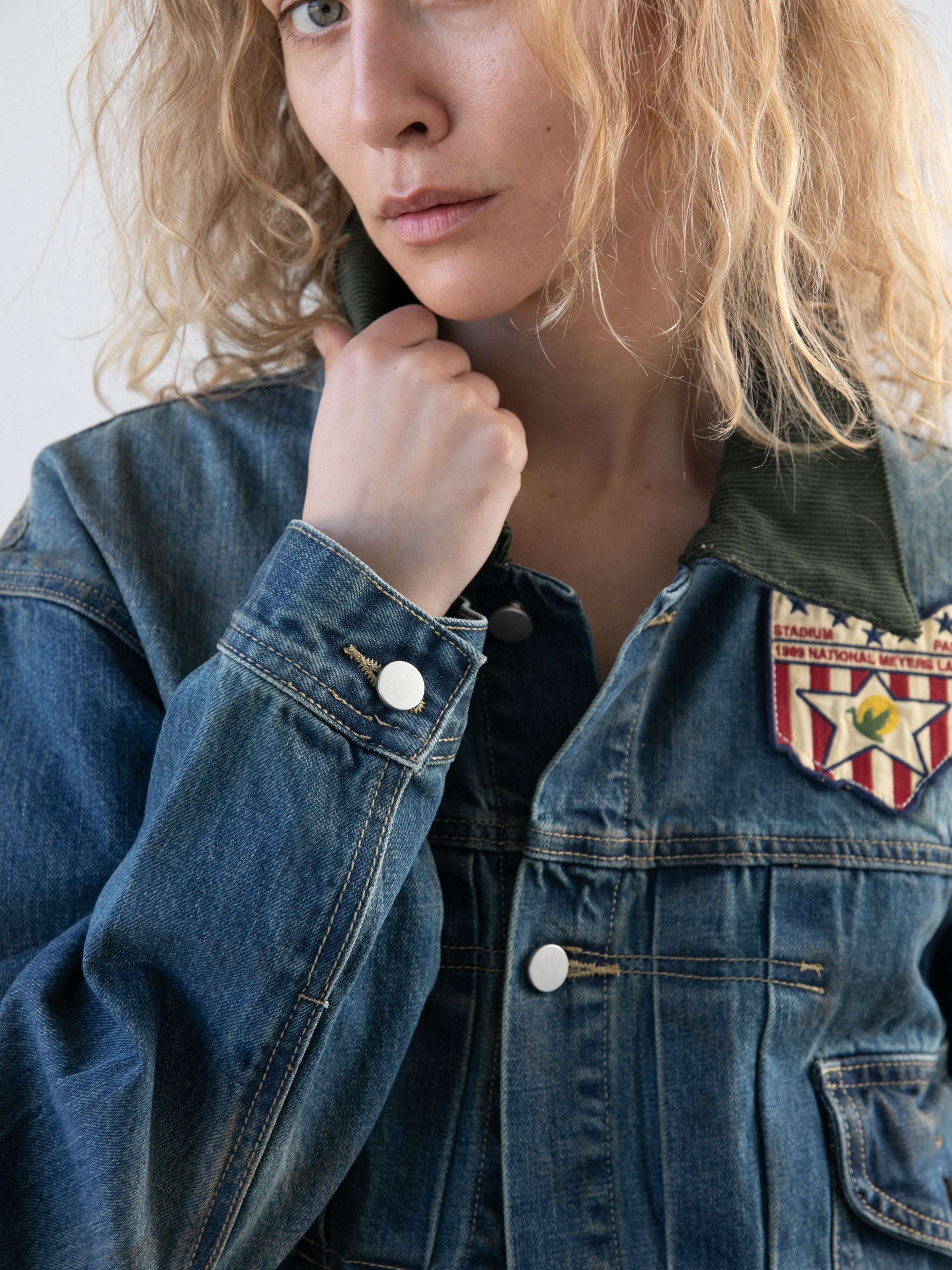 A person with curly blonde hair wears the FOUND Denim Patchwork Jacket, featuring a chest patch and vintage style, while lifting the collar with one hand.