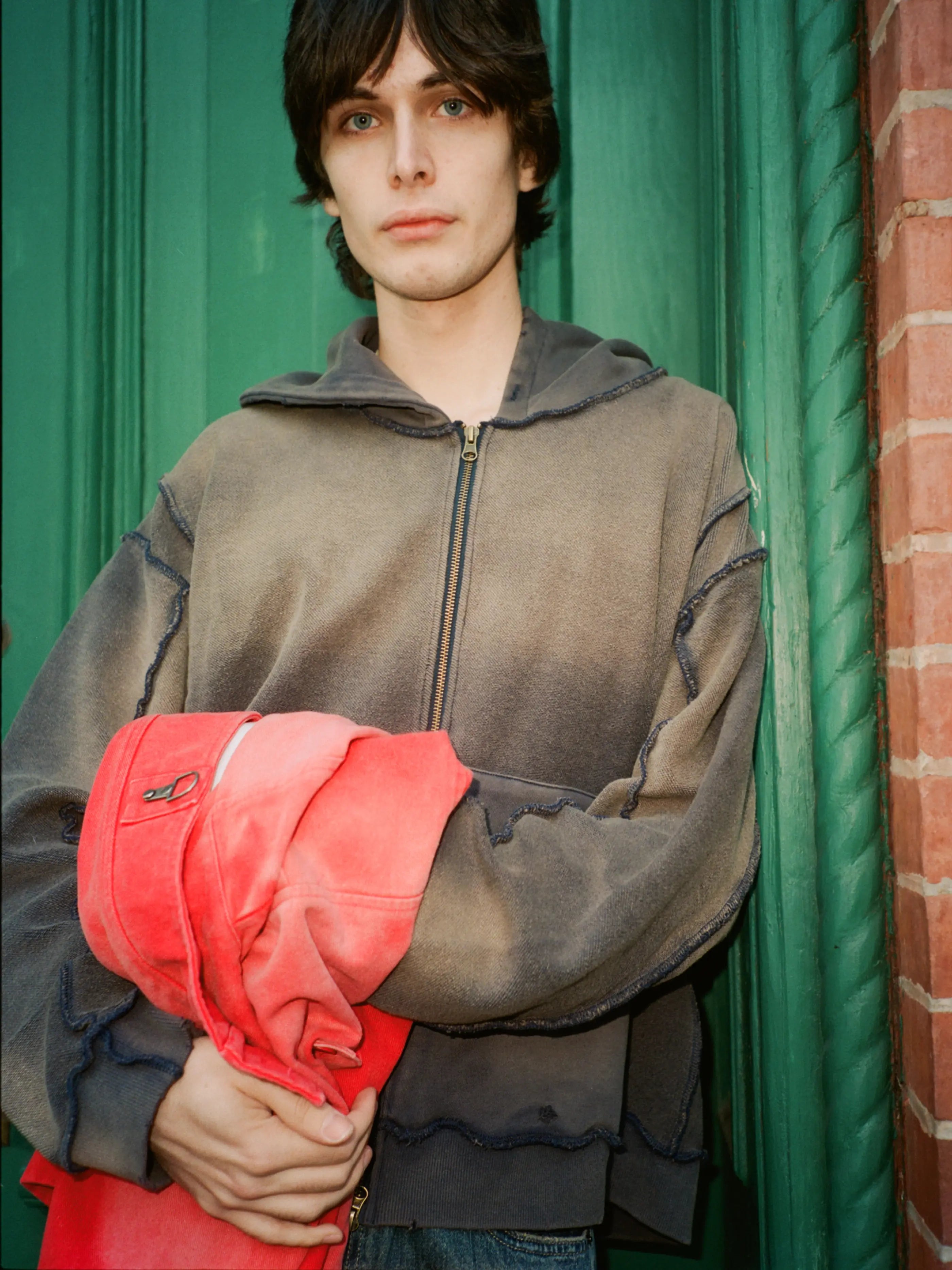 A person stands in front of a green door, wearing the FOUND Deconstructed Terry Zip Up Hoodie and holding a bright red garment.